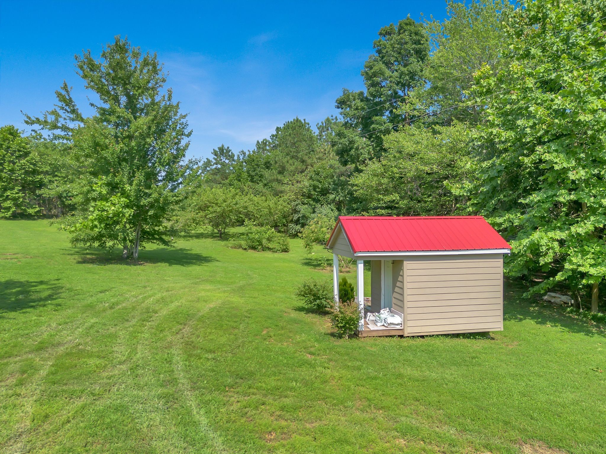 Tan outbuilding with a red metal roof on a broad lawn among mature trees.