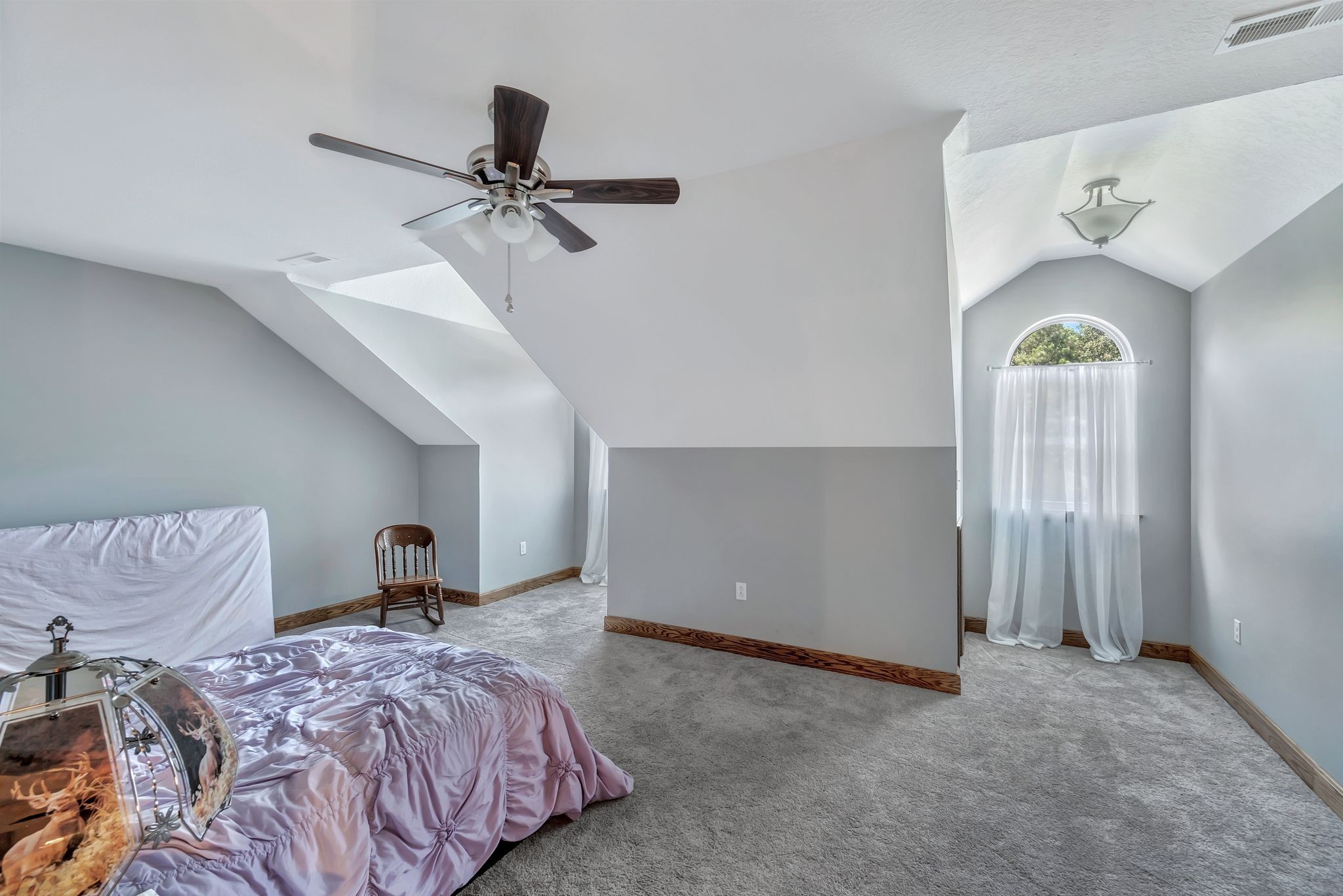 Upstairs bedroom with a lavender-covered bed, sloped ceiling, and arched curtained window.