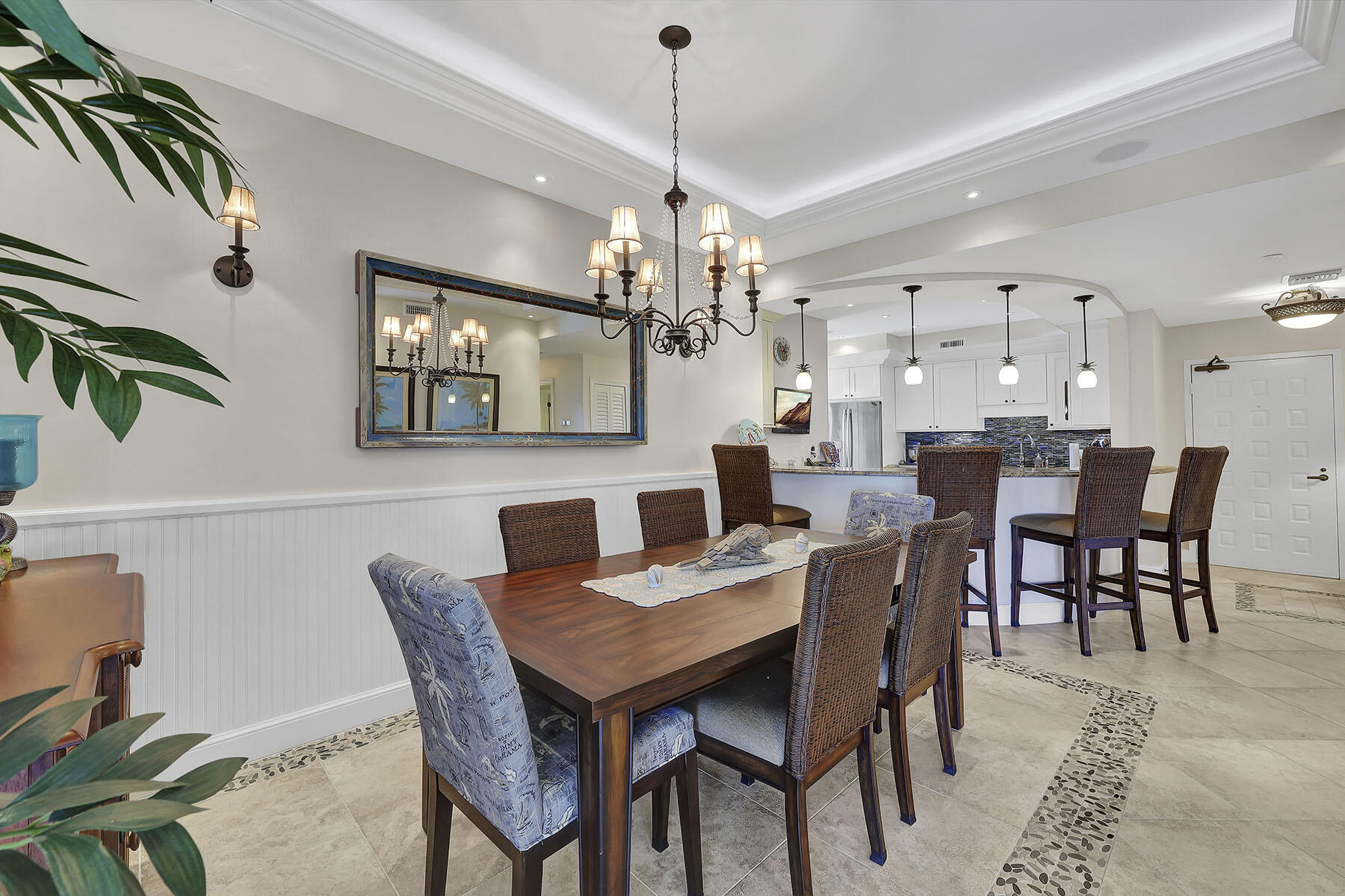 Dining room with a wooden table, woven chairs, chandelier, and open kitchen with blue mosaic backsplash.