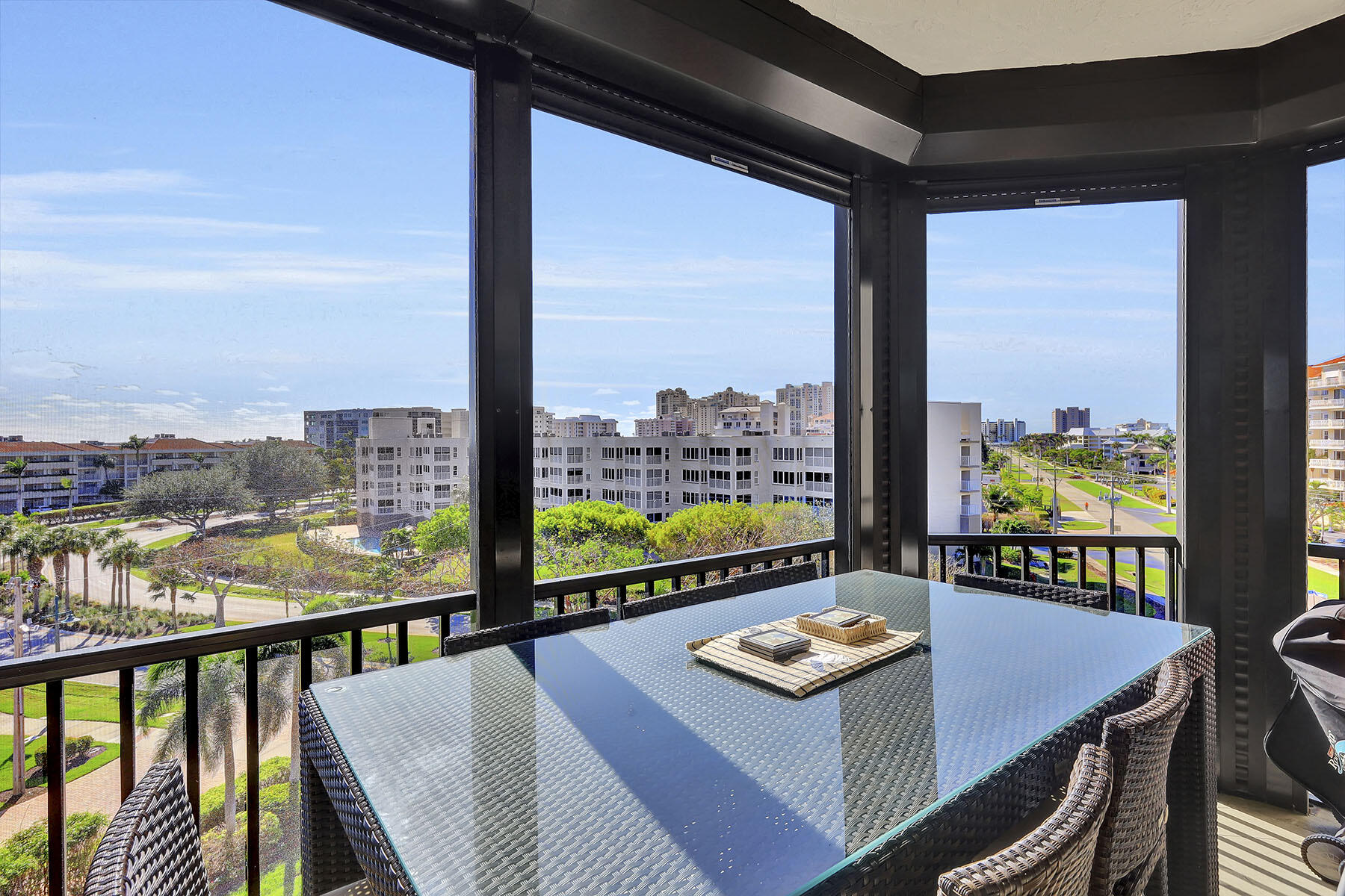 Covered balcony with wicker dining set and views of palm-lined streets and nearby buildings.
