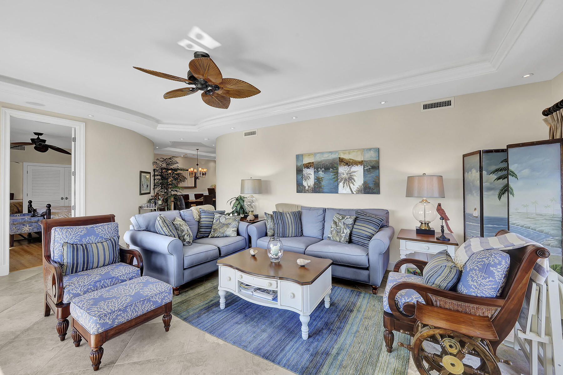 Living room with blue sofas, patterned armchairs, a tropical ceiling fan, and palm-themed artwork.
