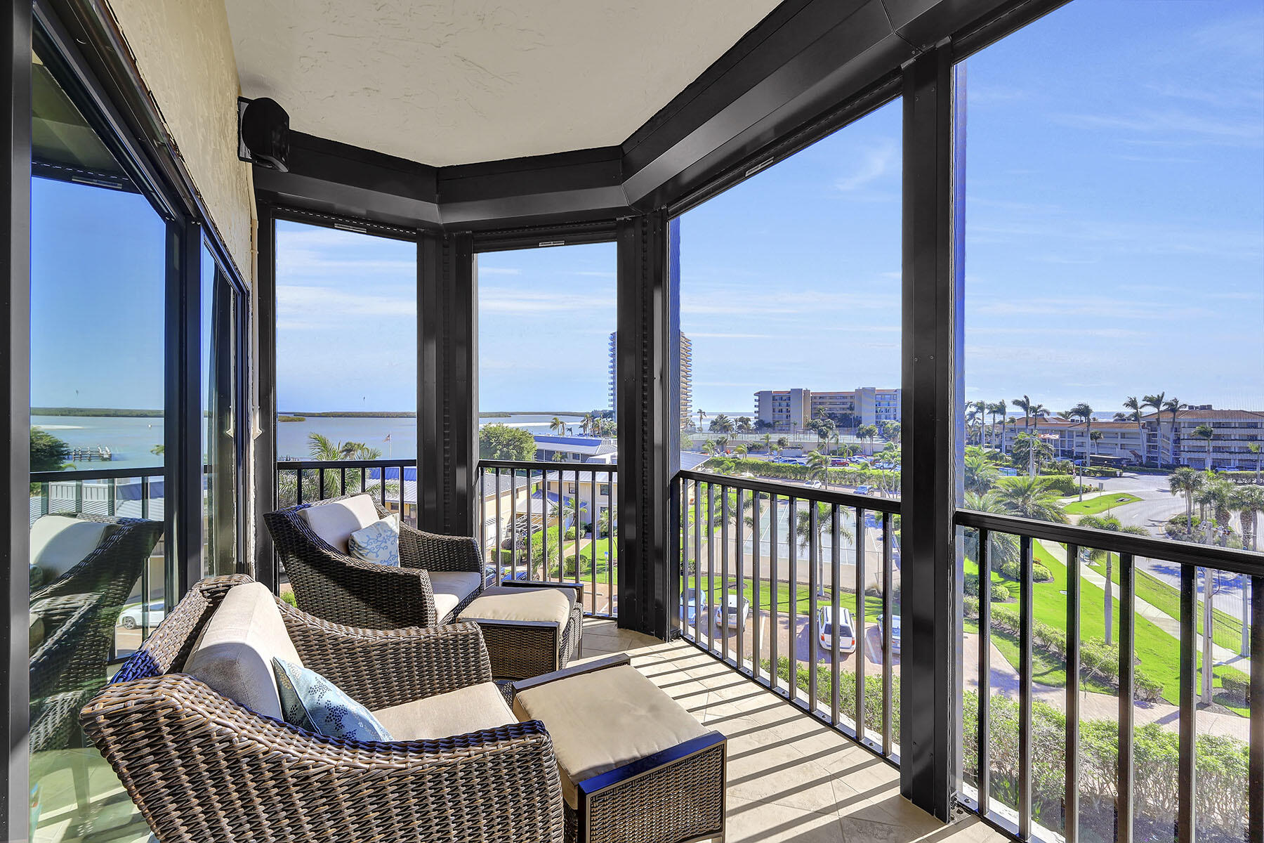 Furnished balcony with wicker seating and coastal water views through black-framed glass panels.