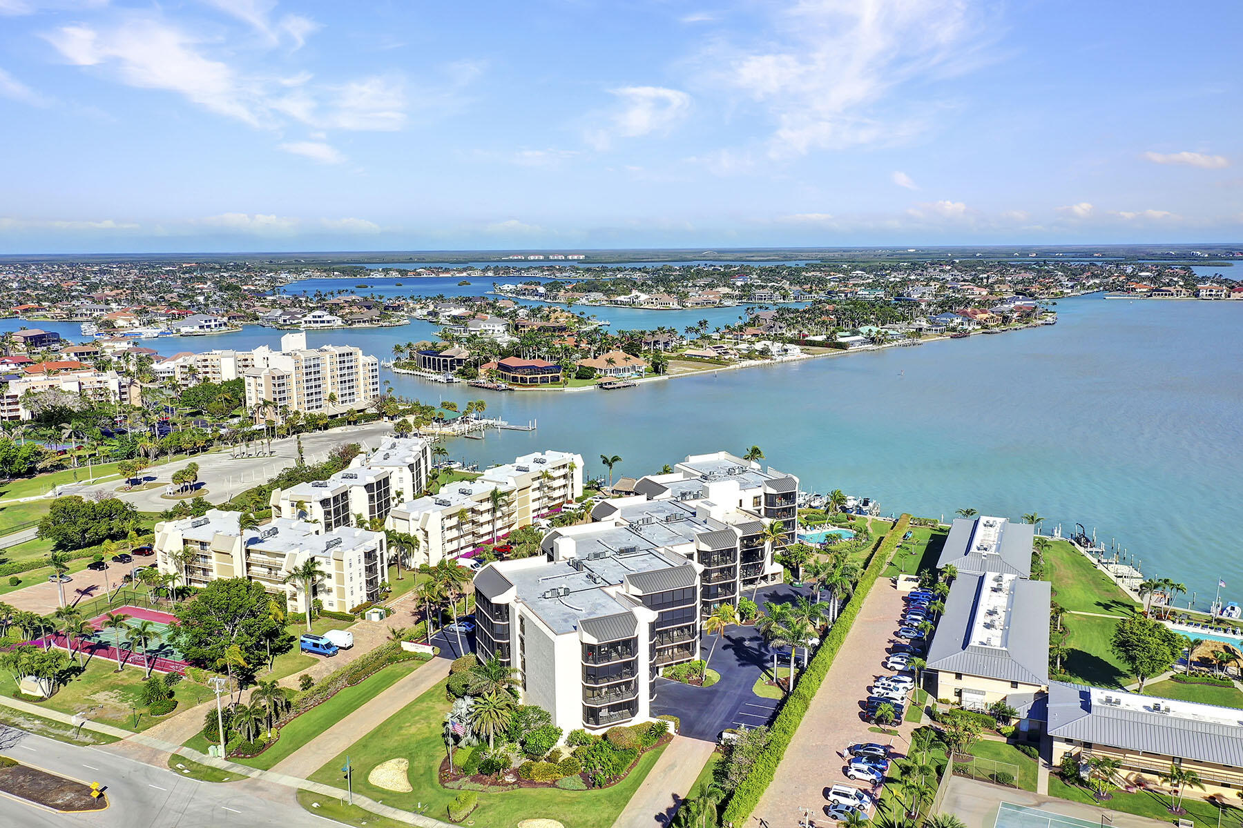 Aerial view of waterfront condominiums beside a broad blue bay, with docks, a pool, and tennis courts.
