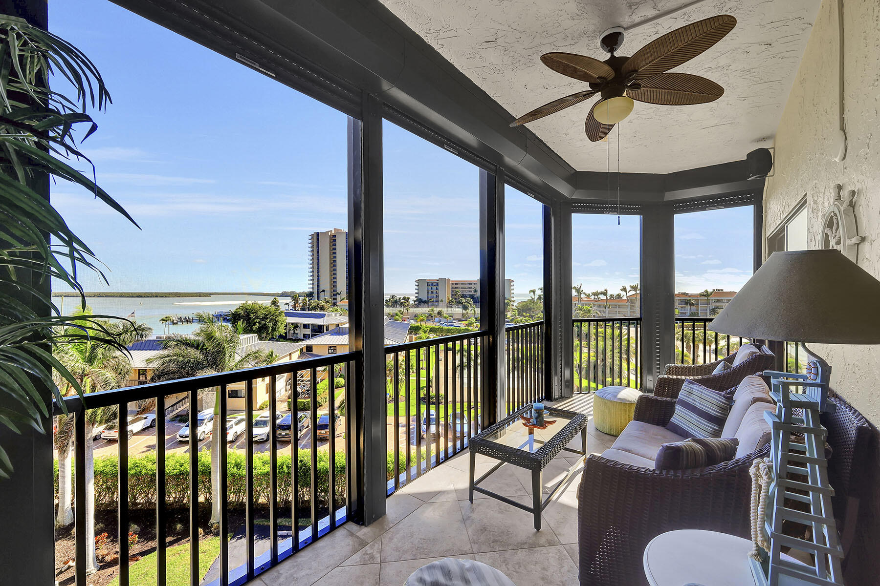 Furnished condo balcony overlooking water and palm-lined grounds, with wicker seating and a ceiling fan.