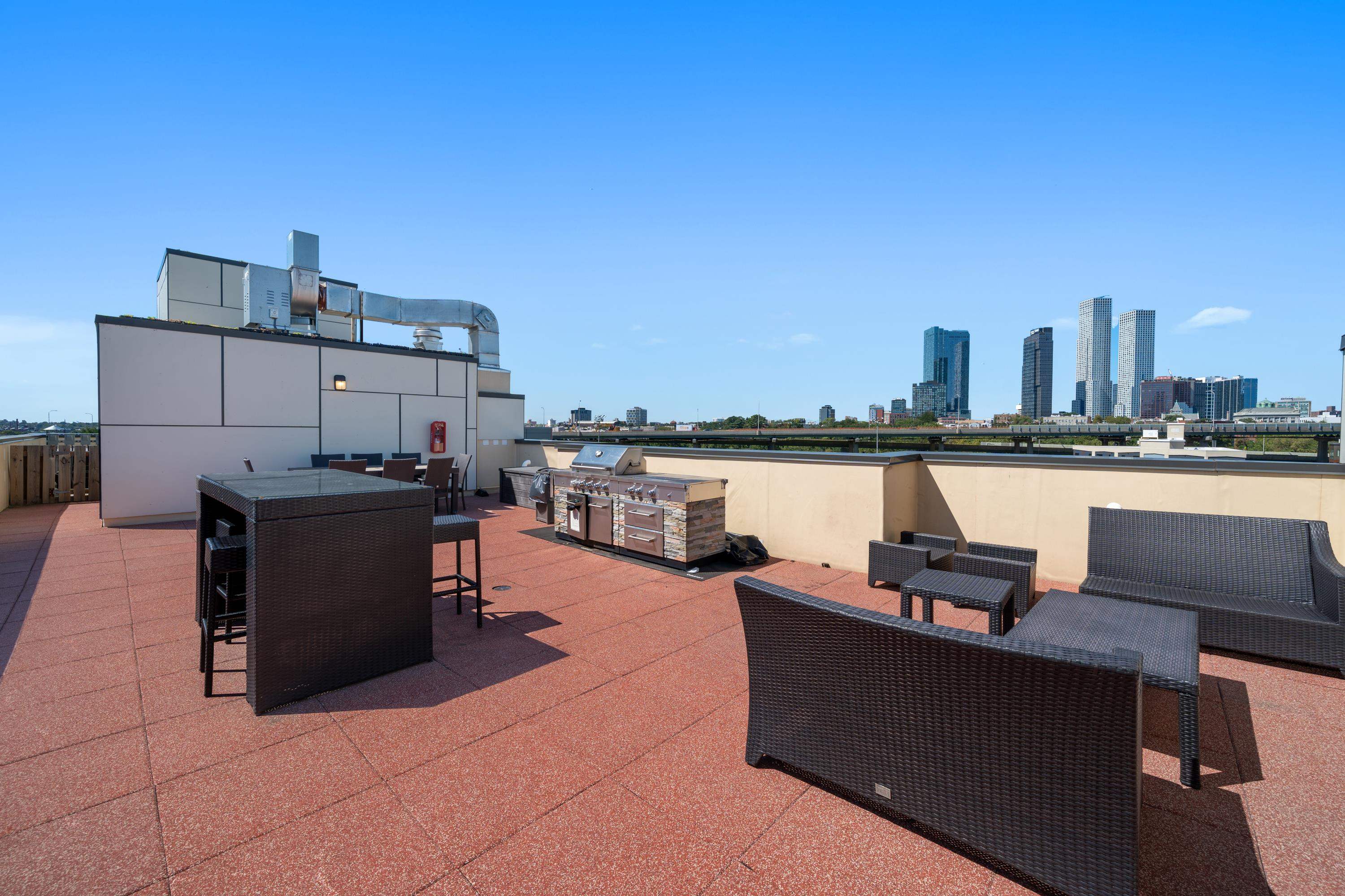 Furnished rooftop terrace with a grill, wicker seating, and a distant city skyline.