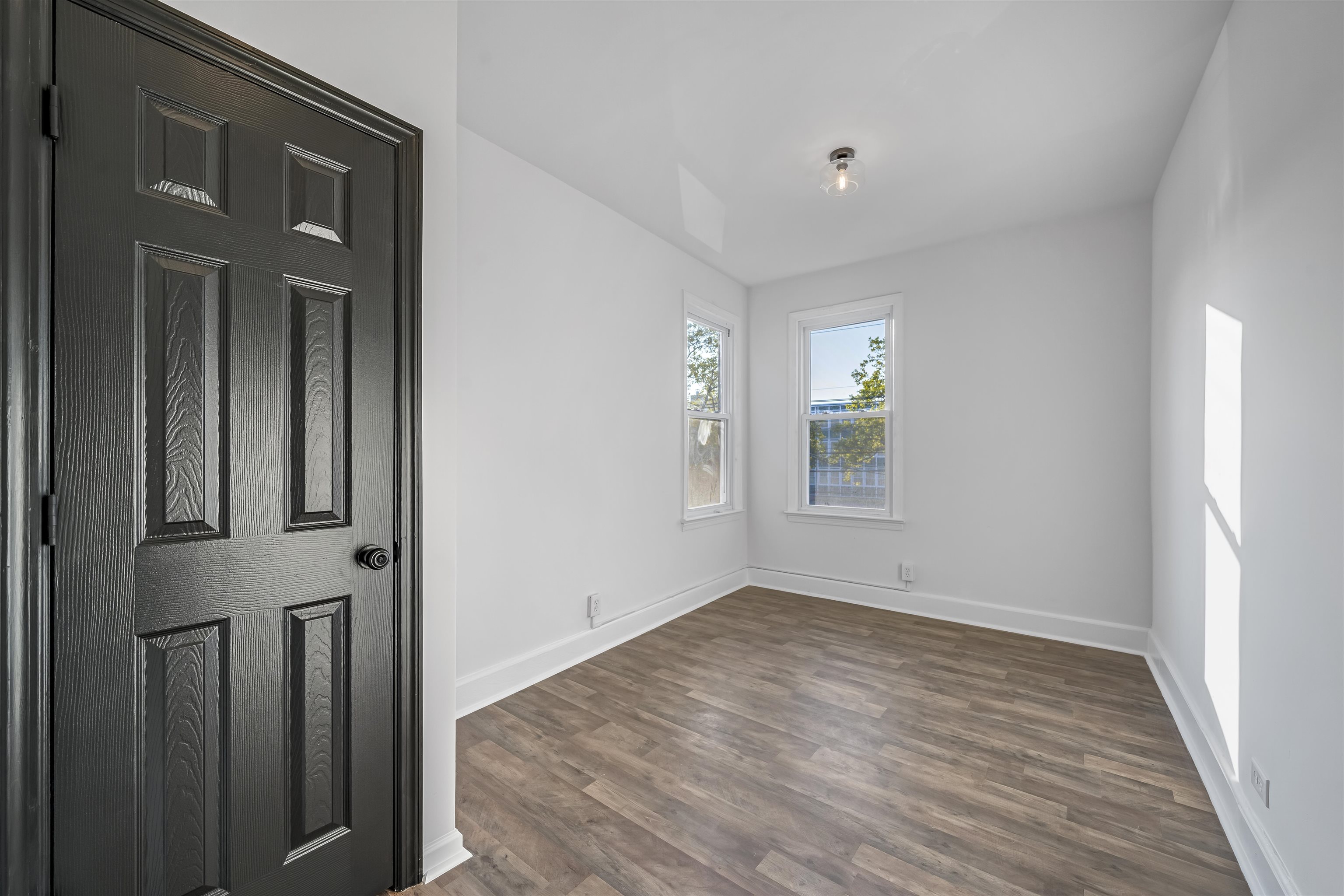 Empty bedroom with two windows, wood-look flooring, and a dark paneled door.