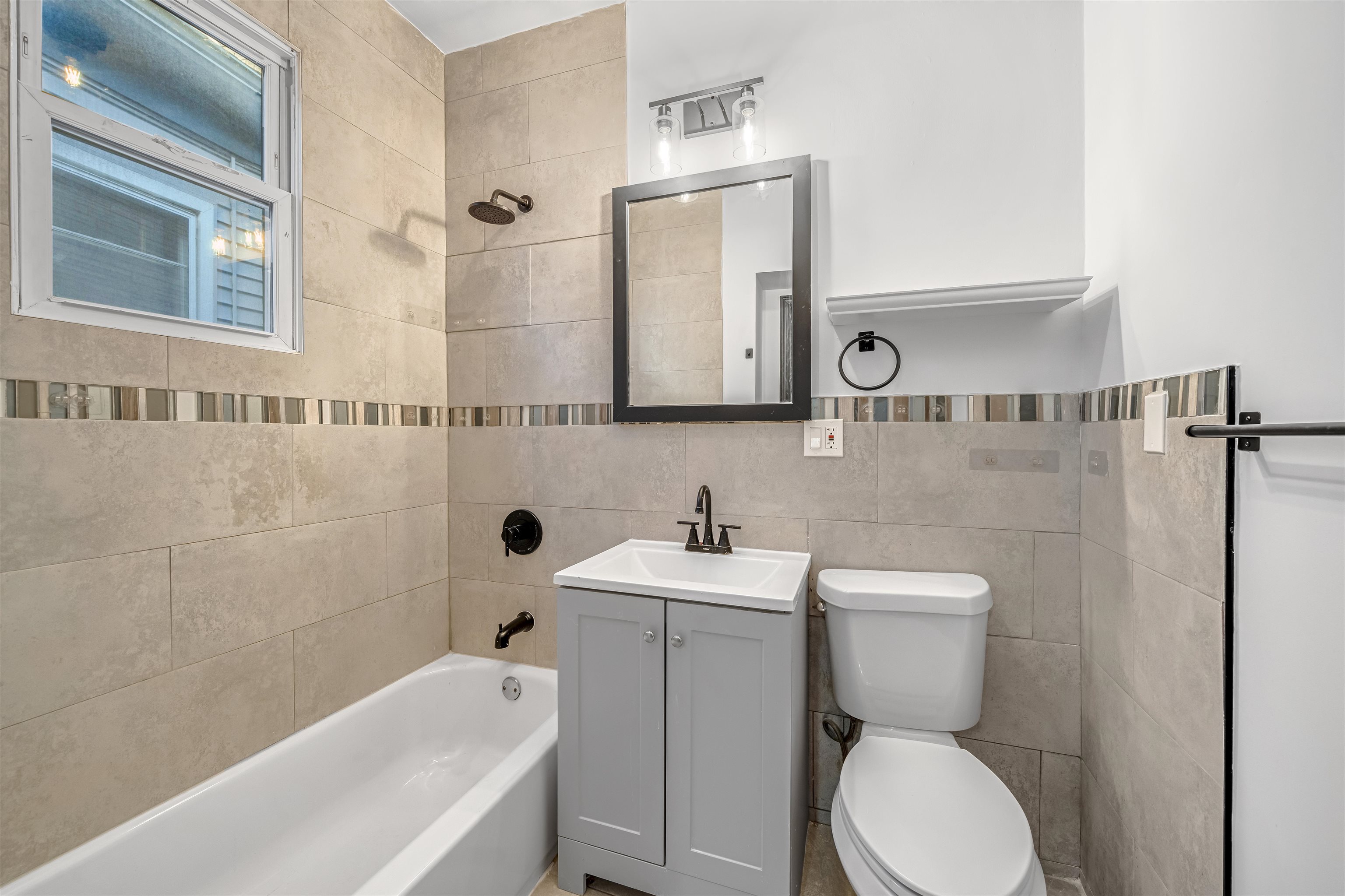 Bathroom with beige tile, a bathtub beneath a window, and a gray vanity beside the toilet.