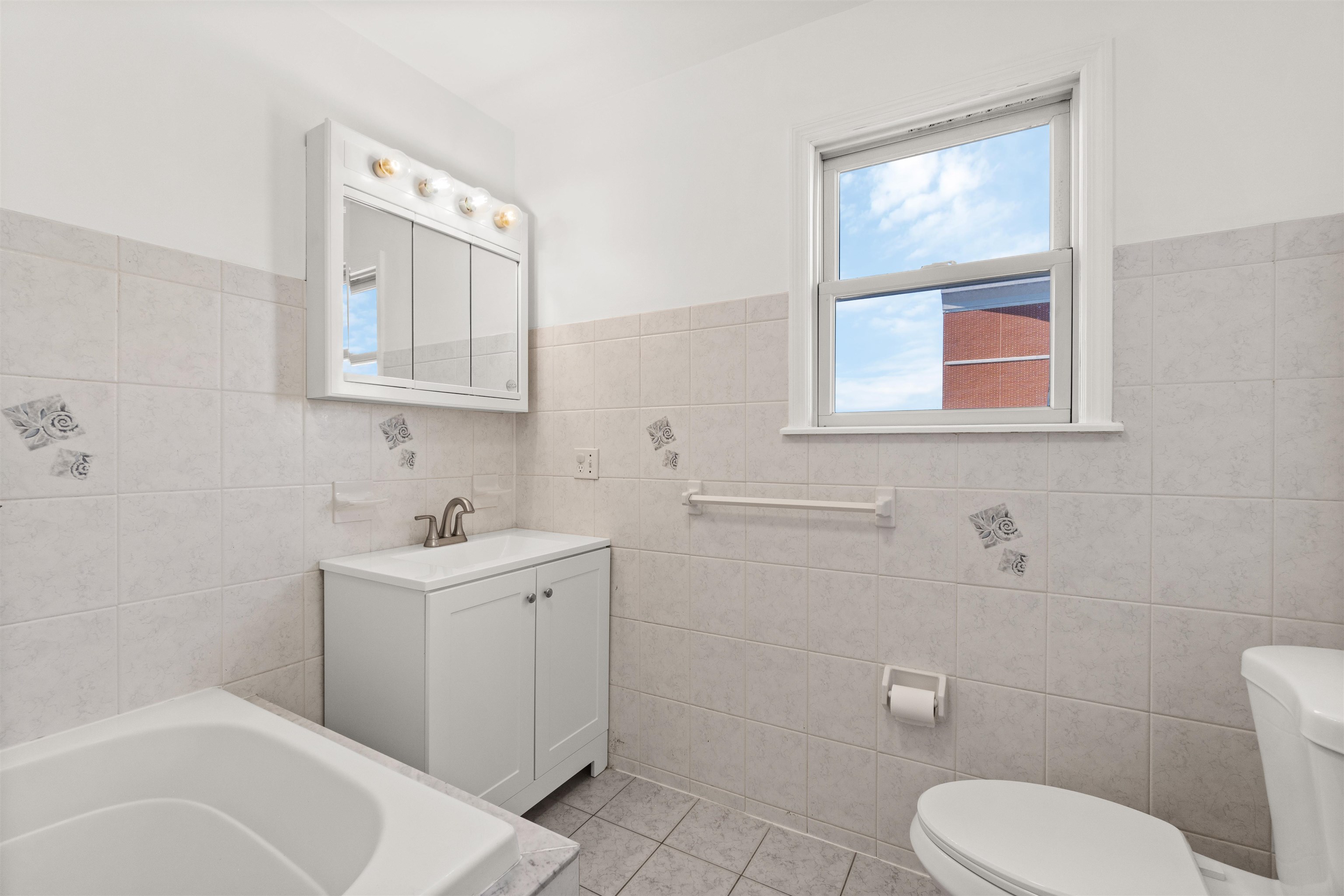 Tiled bathroom with a white vanity, mirrored medicine cabinet, bathtub, and window.
