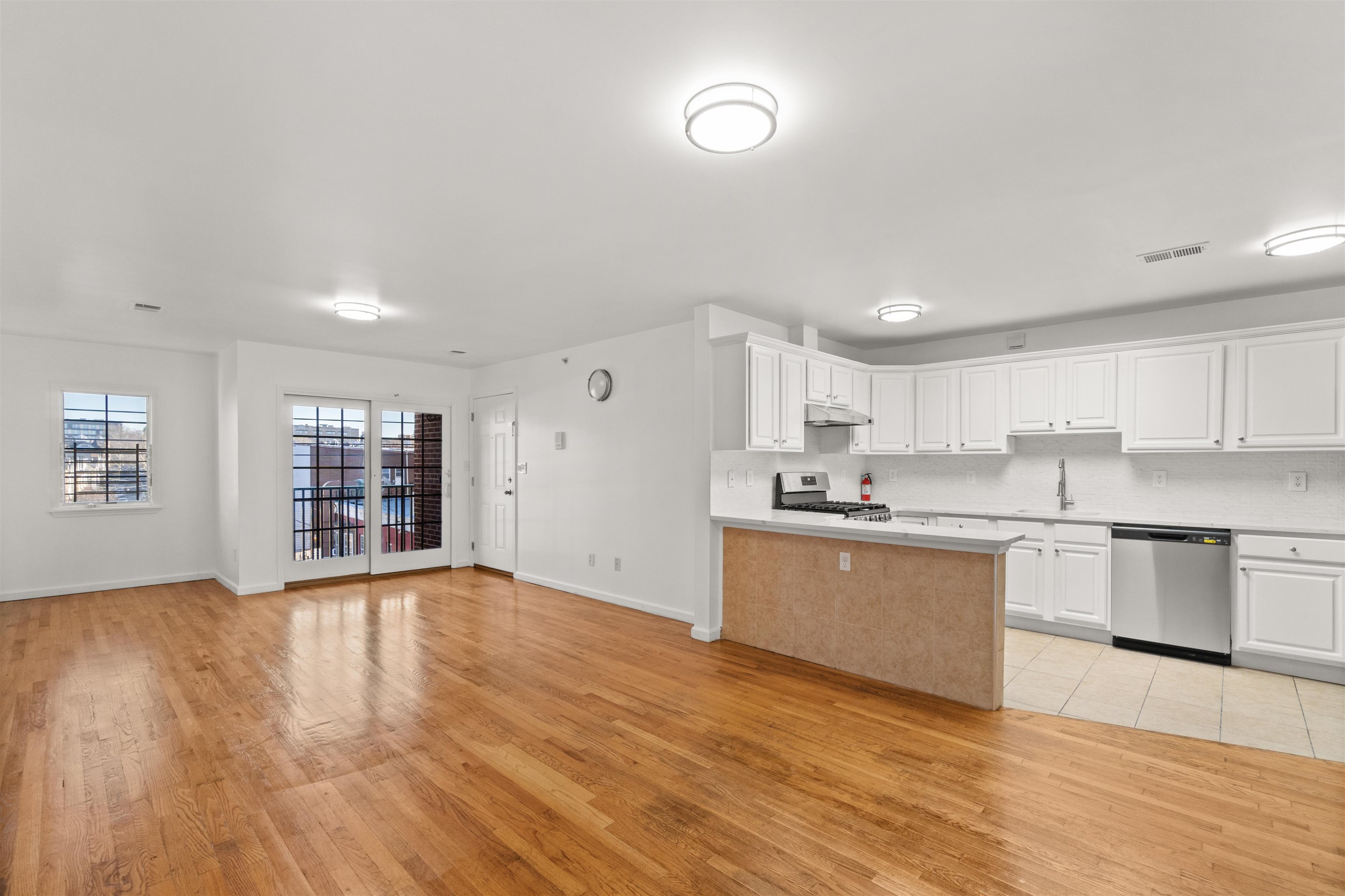 Open-plan apartment kitchen and living area with white cabinets, a tiled peninsula, hardwood floors, and balcony doors.