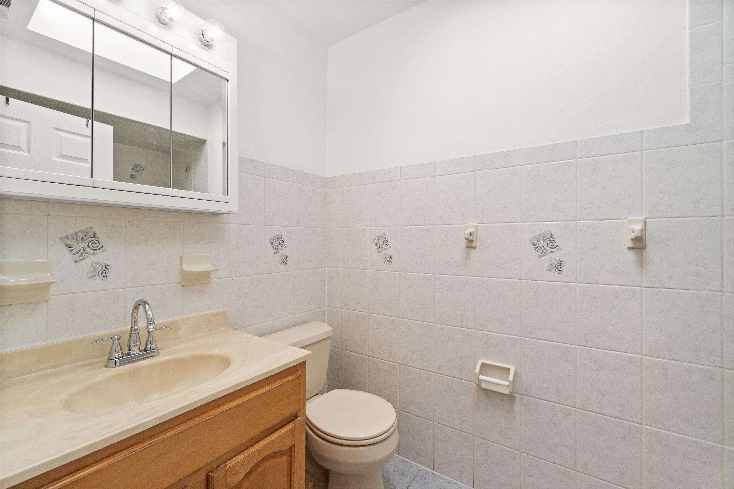 Bathroom with a wood vanity, mirrored medicine cabinet, toilet, and cream tile with blue accents.
