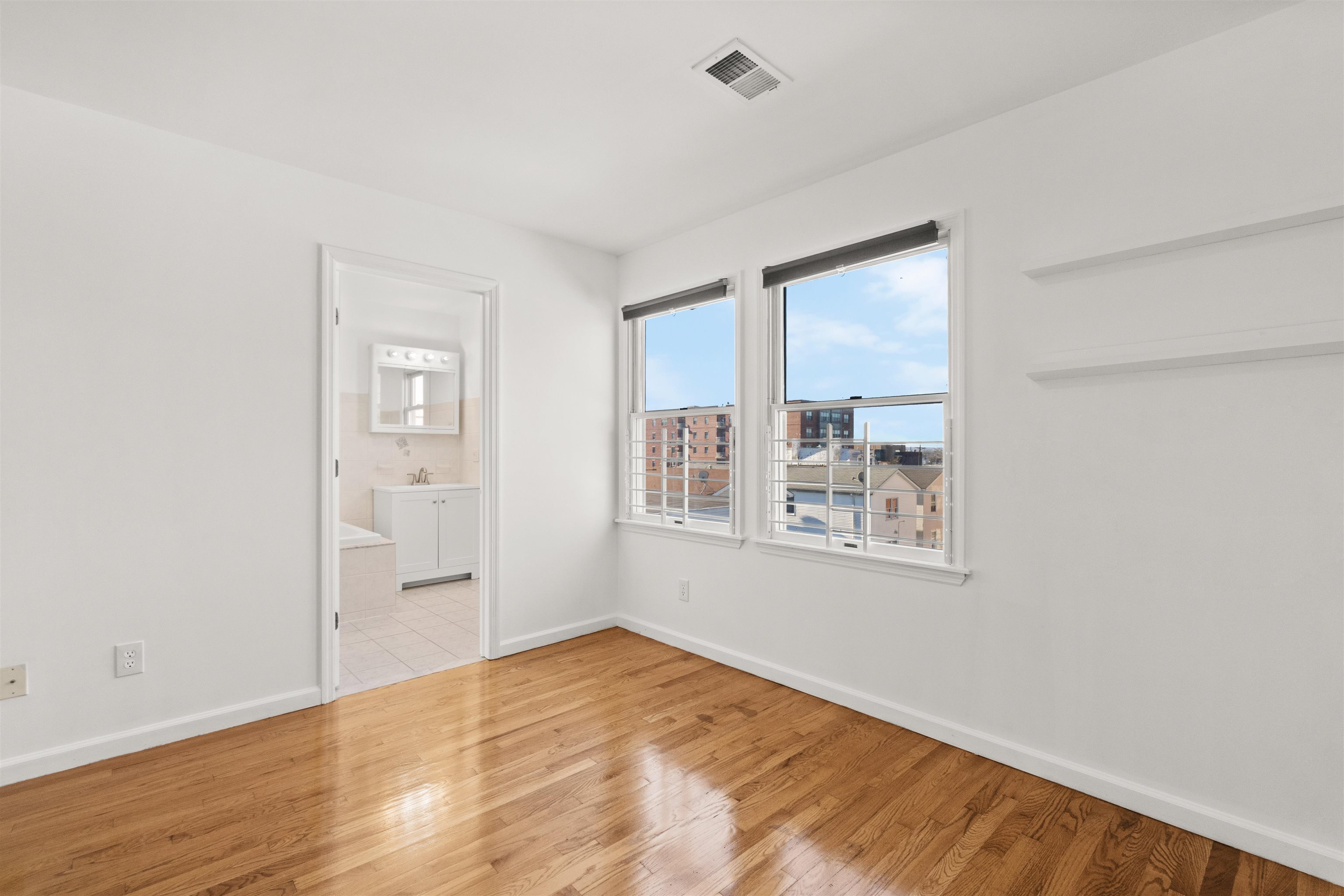 Empty room with polished hardwood floors, two windows, and a bathroom doorway.