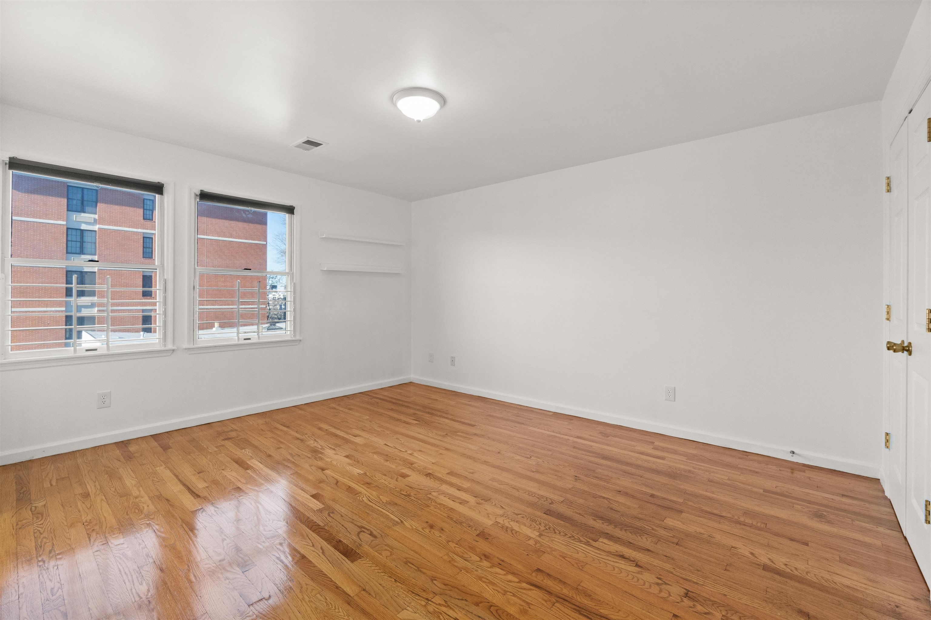 Empty room with polished hardwood floors, two windows, and white wall shelves.