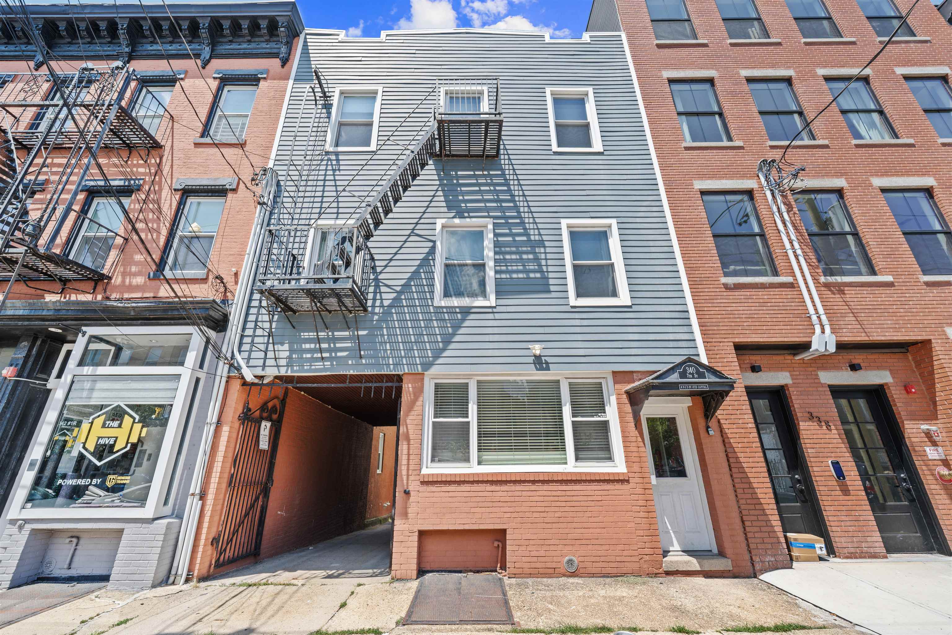 Narrow gray-sided building with a metal fire escape and brick entrance between taller brick buildings.