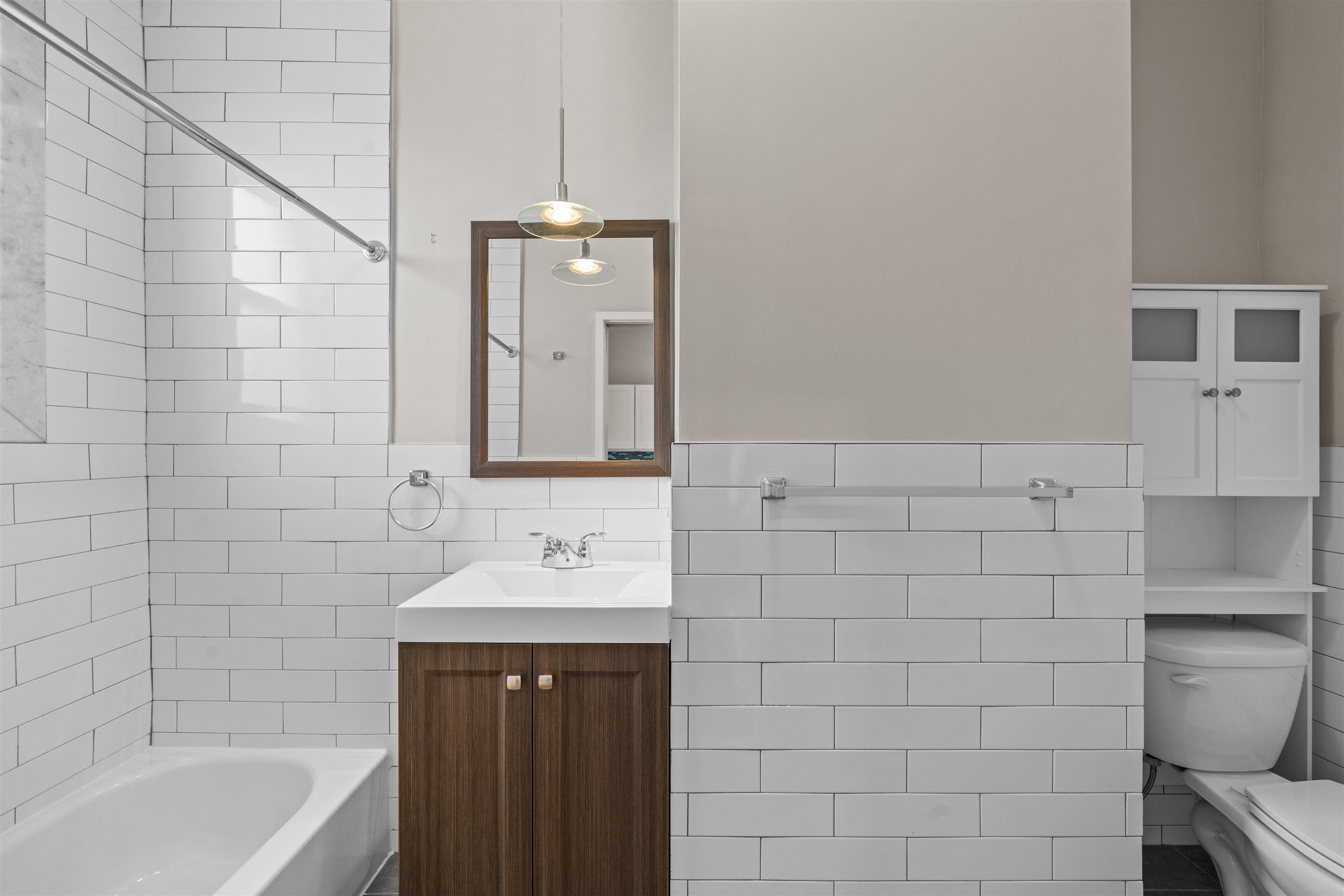 Bathroom with white subway tile, a dark wood vanity, bathtub and toilet behind a half wall.