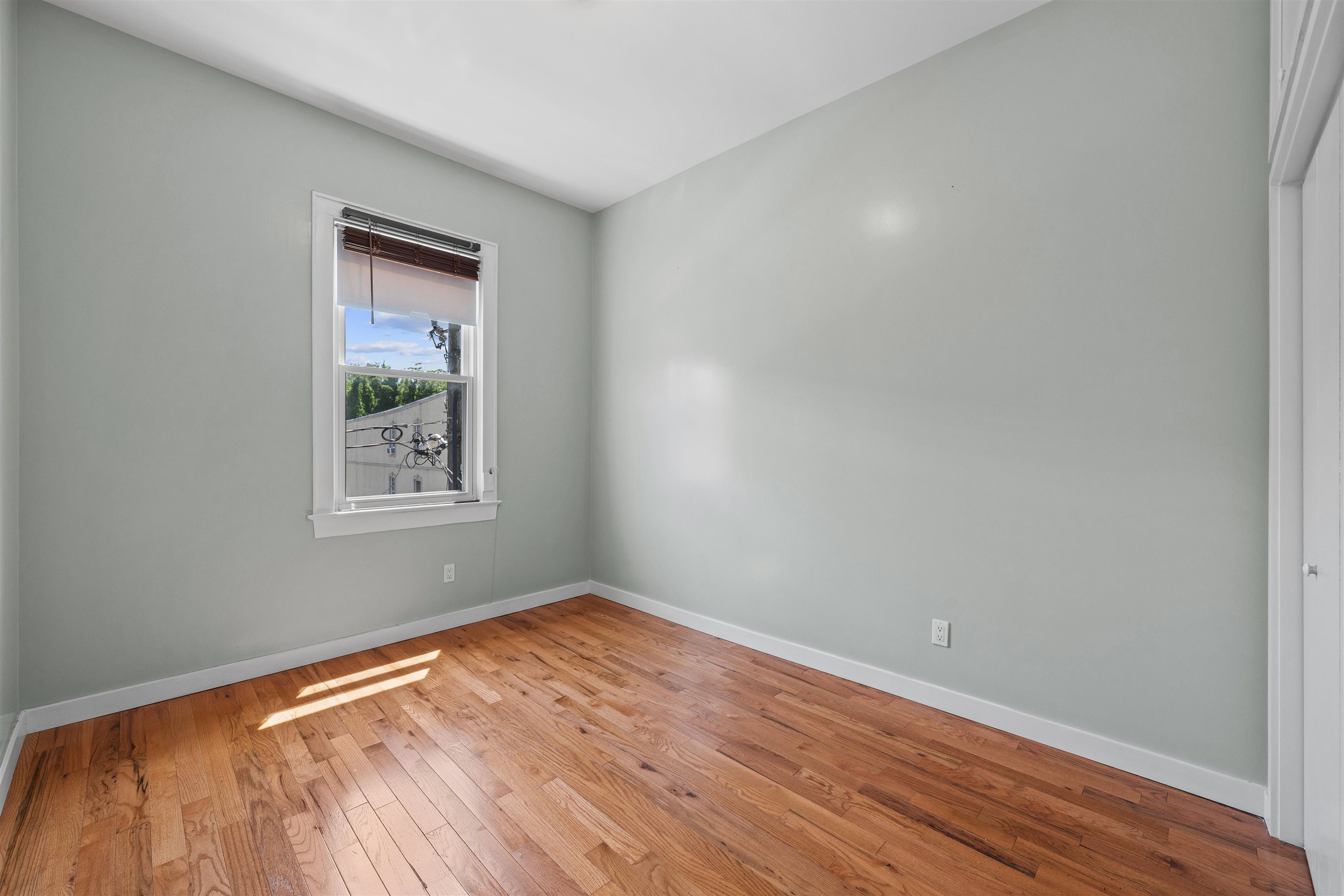Empty bedroom with hardwood floors, pale gray walls, and a sunlit window with blinds.