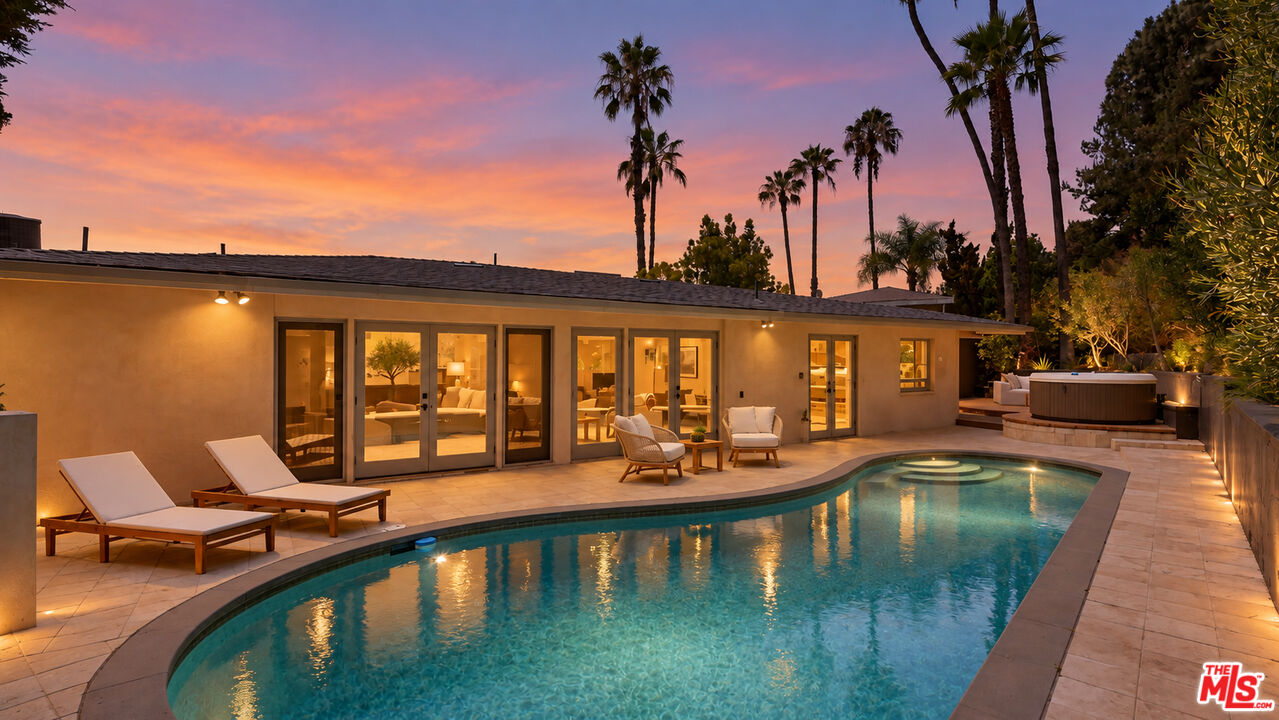 Single-story home beside a curved pool and raised hot tub at dusk, with palm trees against a pink sky.