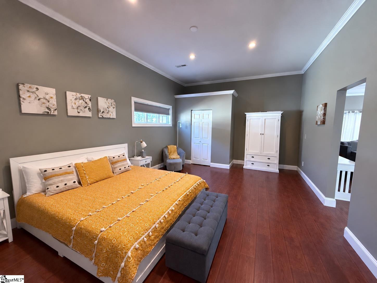 Staged bedroom with a mustard-yellow bedcover, gray bench, floral art, and white wardrobe.