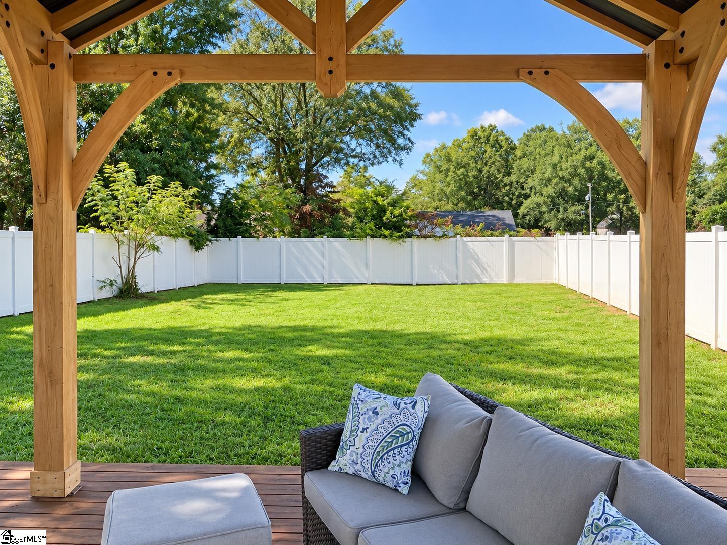 Wooden pergola patio with gray wicker seating overlooking a fenced green backyard.