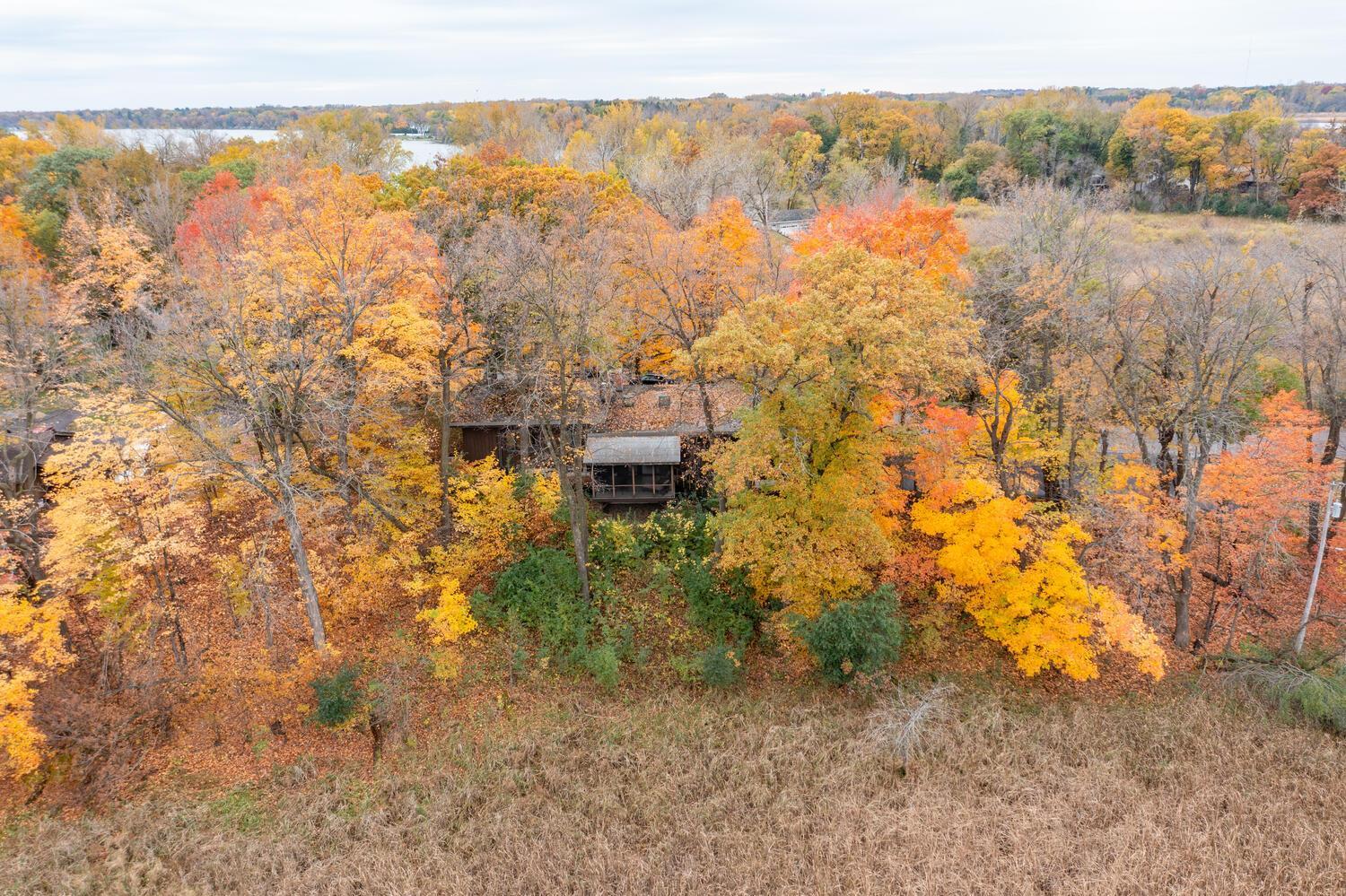 Aerial view of a wooded home surrounded by vivid autumn trees, with water visible beyond.
