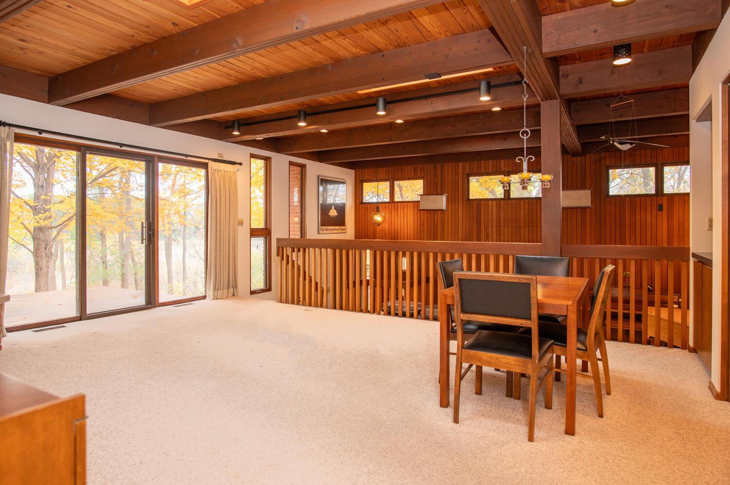 Dining area with exposed wood beams, a wooden table, and sliding glass doors overlooking autumn trees.