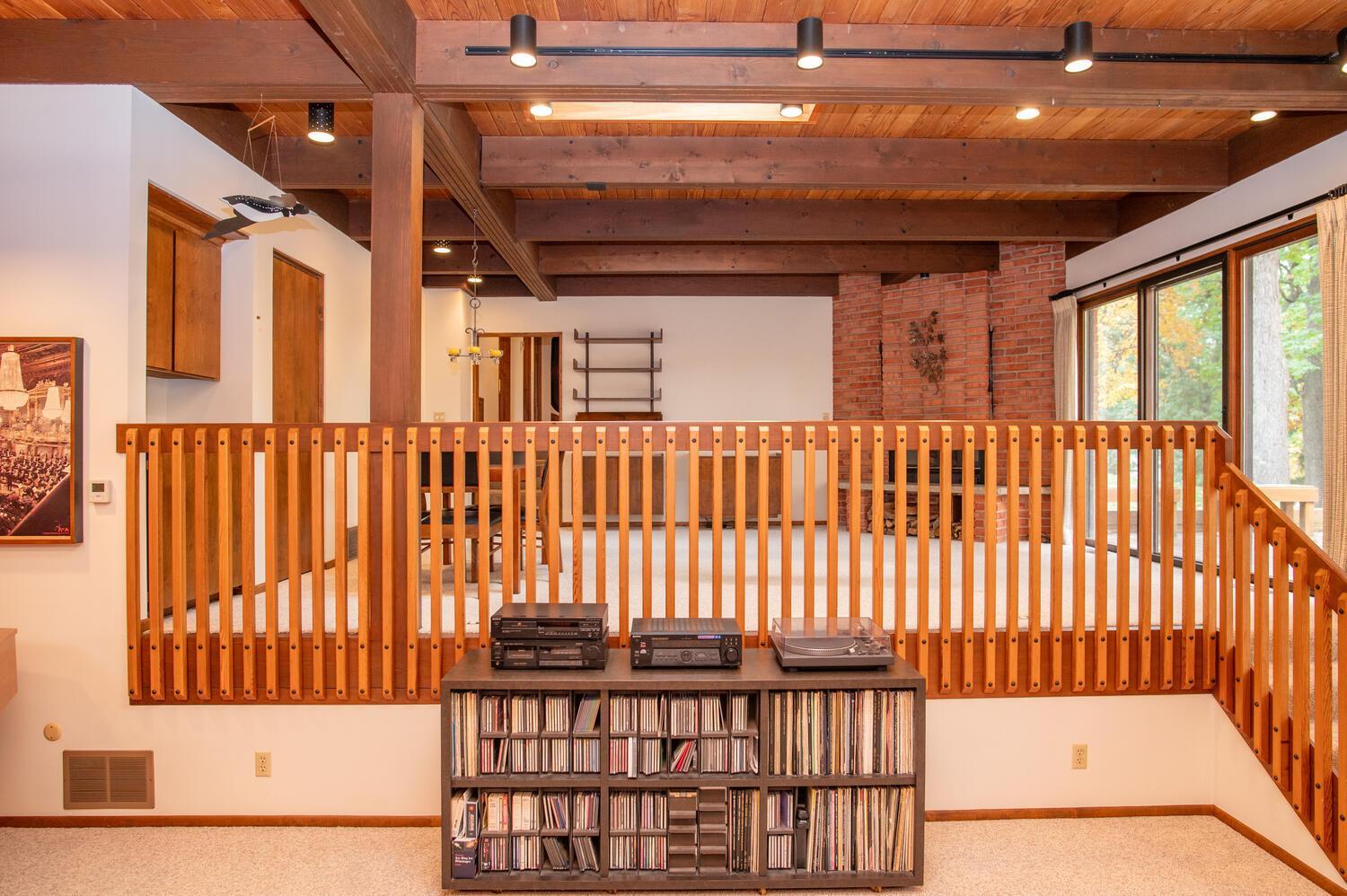 Living room with exposed wood beams, brick fireplace, large windows, and a wooden railing.