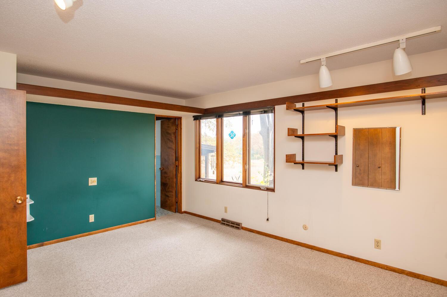 Empty carpeted room with a teal accent wall, large window, and built-in wooden shelves.