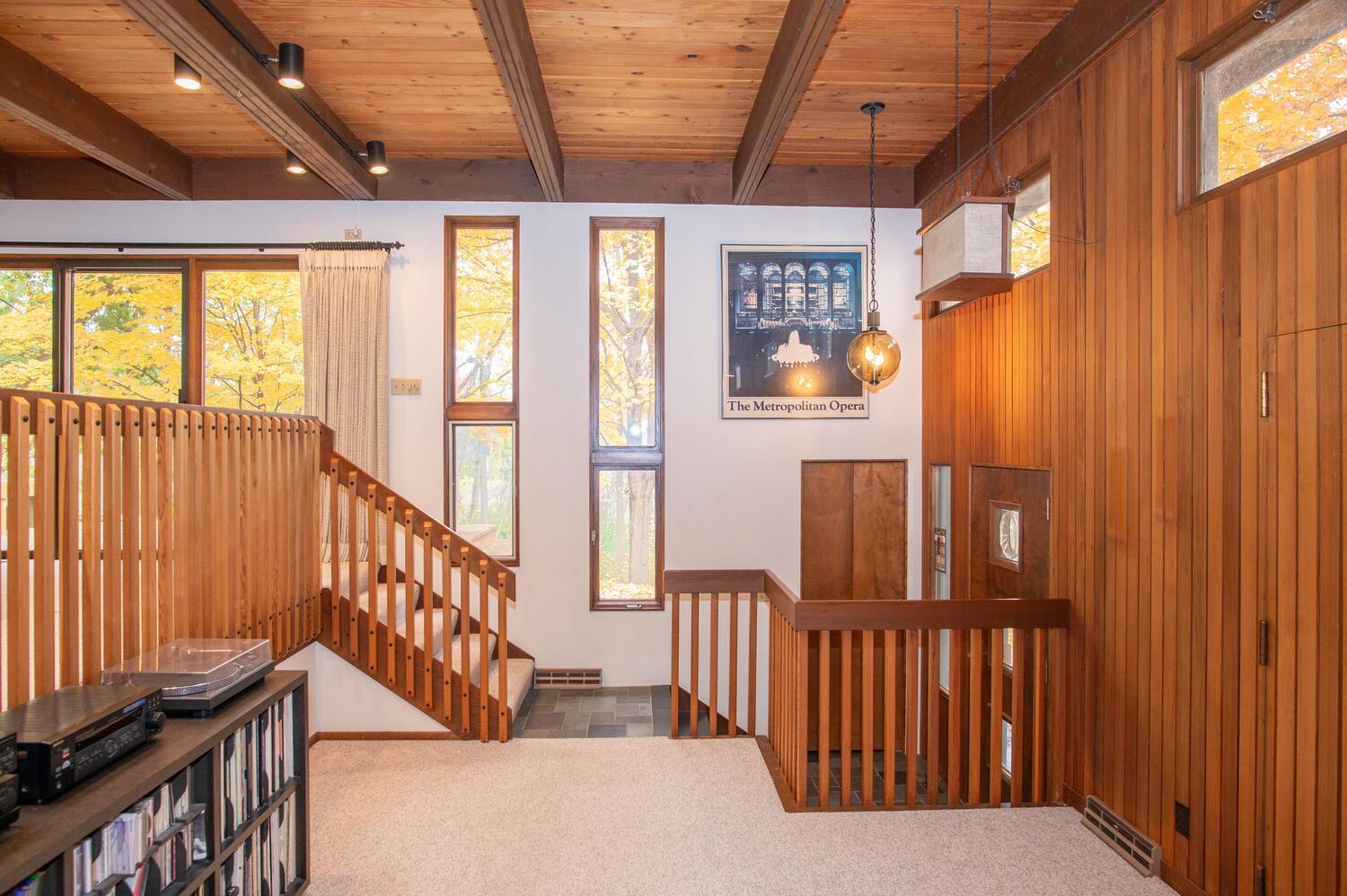 Wood-paneled split-level landing with exposed beams, stair railings, and tall windows facing autumn trees.