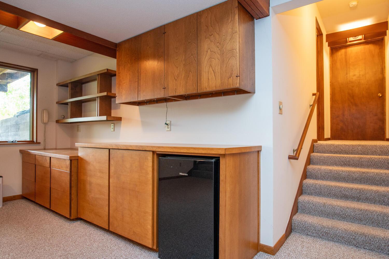 Wood-cabinet kitchenette with a compact refrigerator beside carpeted stairs.