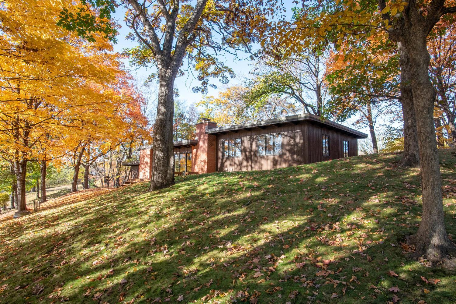 Single-story wood-and-brick home on a grassy slope beneath colorful autumn trees.