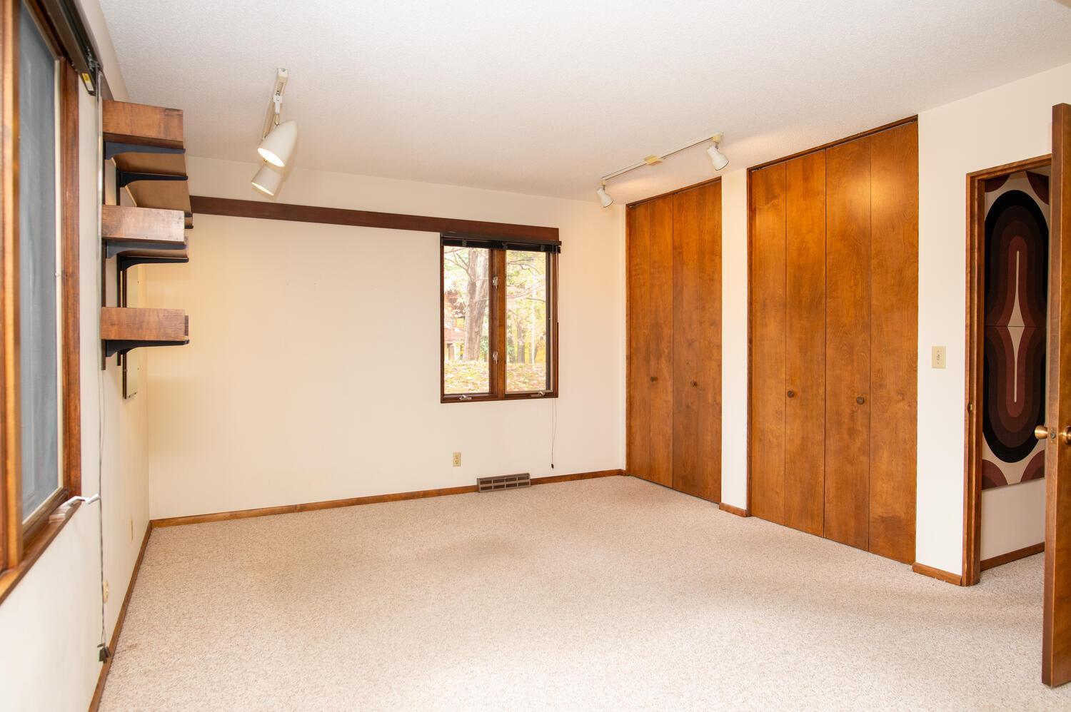 Empty bedroom with beige carpet, built-in wooden closets, and a window overlooking trees.