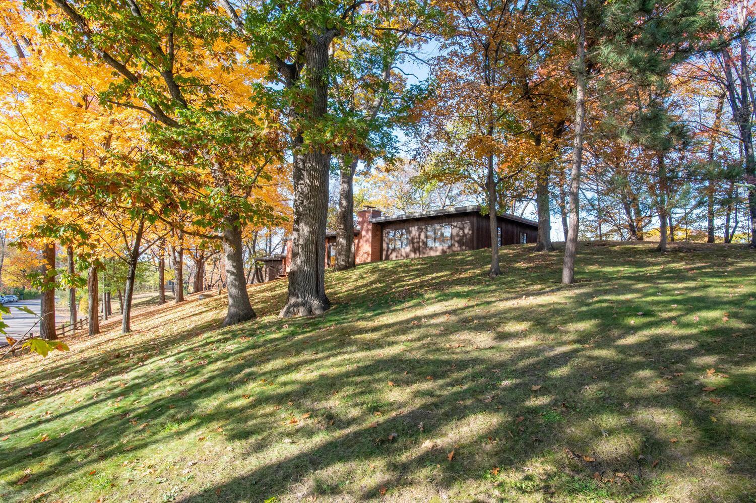 Low brown home on a grassy, wooded slope beneath colorful autumn trees.
