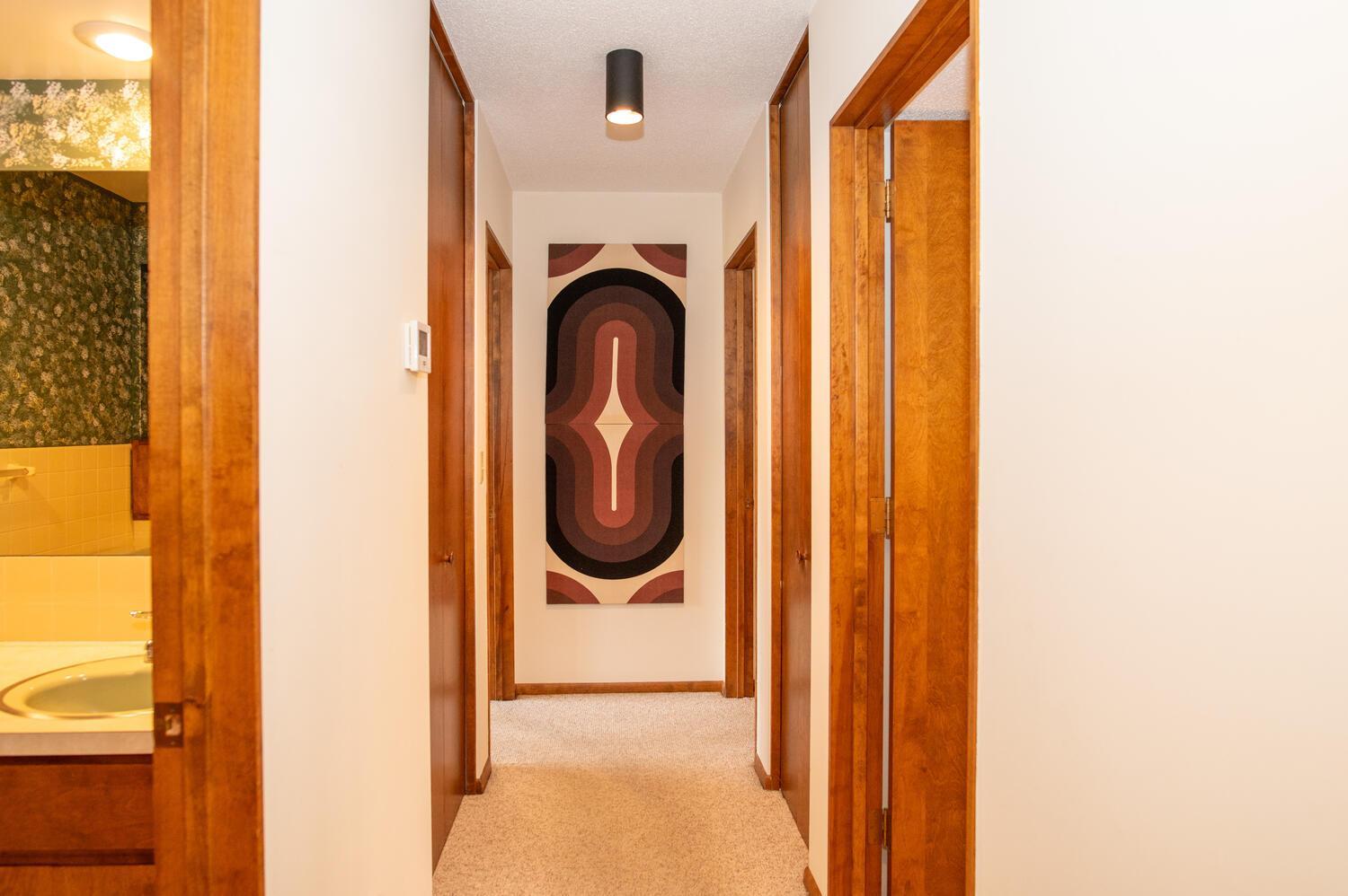 Wood-trimmed hallway with beige carpeting and geometric wall art, viewed past an open bathroom.
