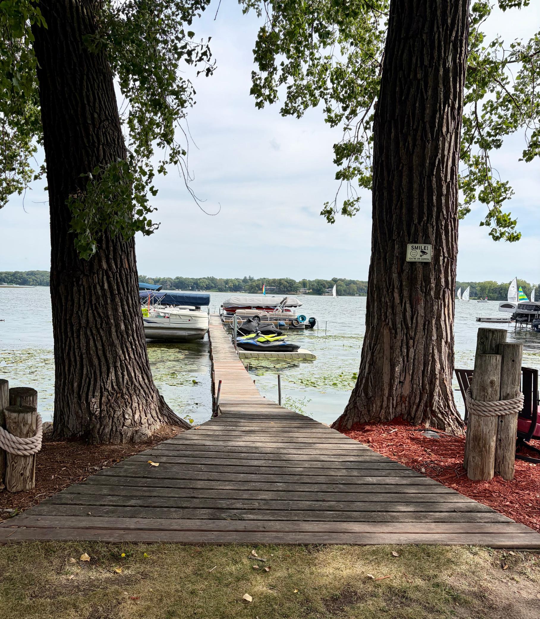 Wooden dock between large trees extends to boats on a lake, with sailboats in the distance.