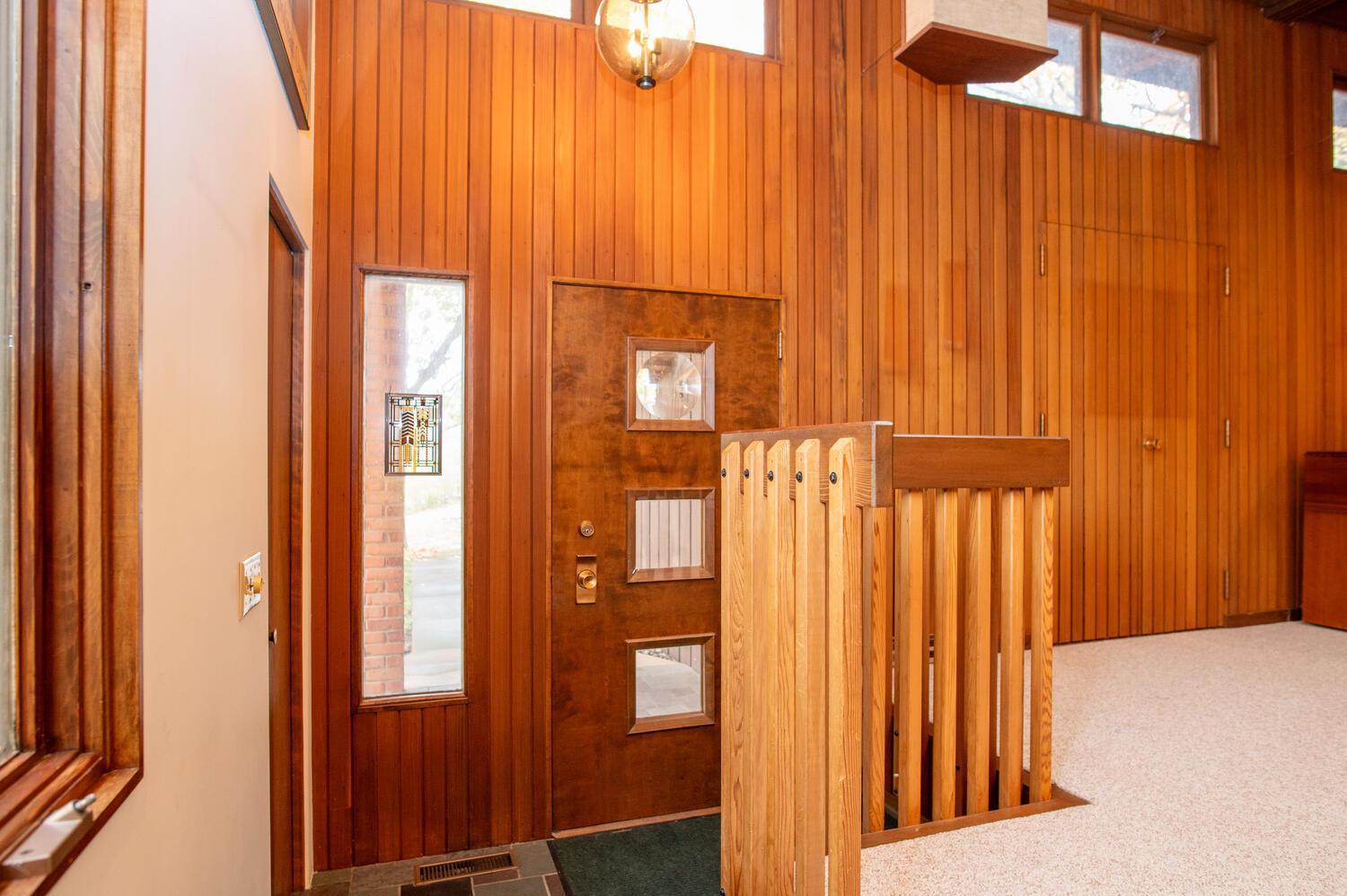 Wood-paneled entryway with a dark front door, wooden stair railing, and high windows.