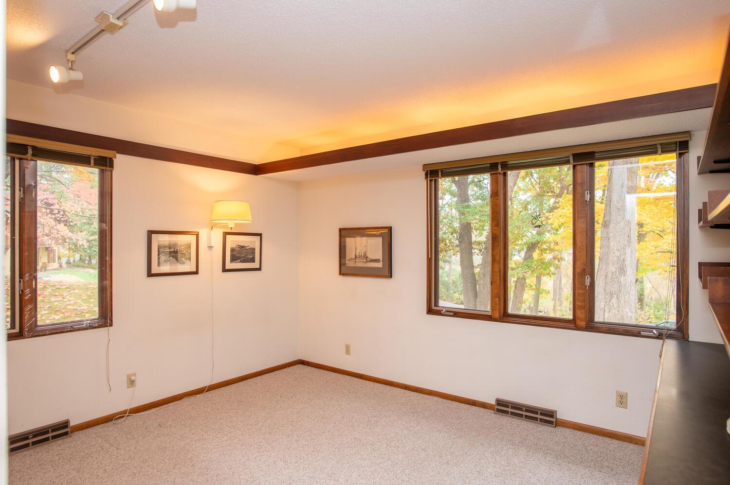 Empty carpeted room with dark wood trim and wide windows overlooking autumn trees.