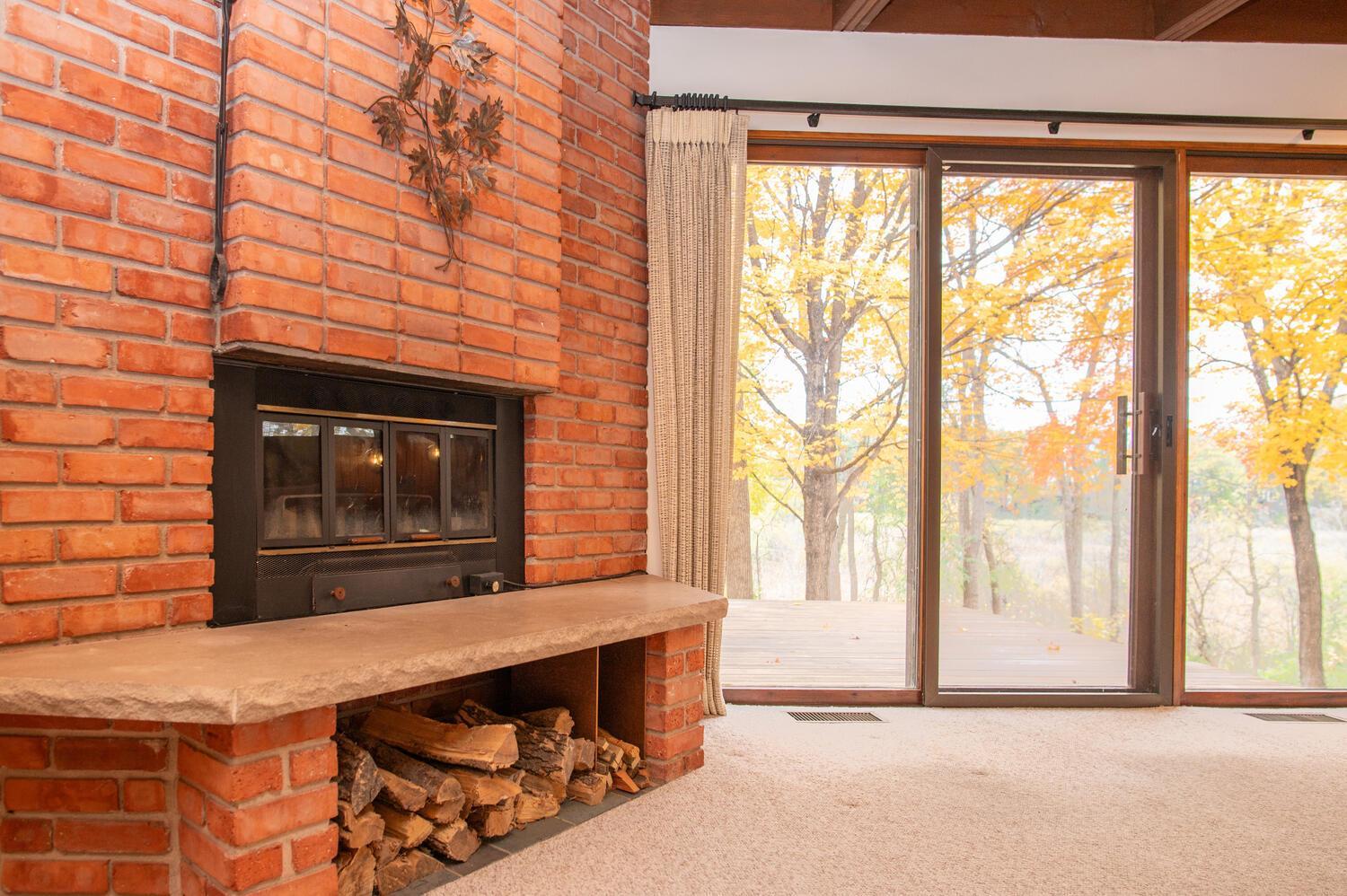 Living room with a red-brick fireplace and sliding glass doors overlooking autumn trees.