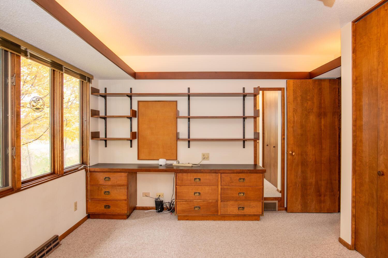 Home office with built-in wooden desk and shelves beside windows overlooking autumn foliage.