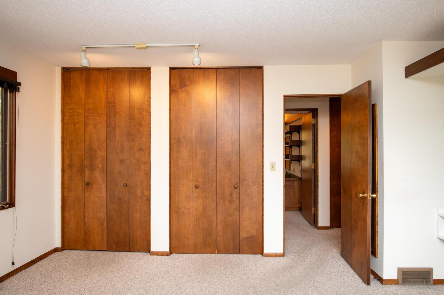 Empty bedroom with beige carpet, wooden closets, and an open bathroom doorway.