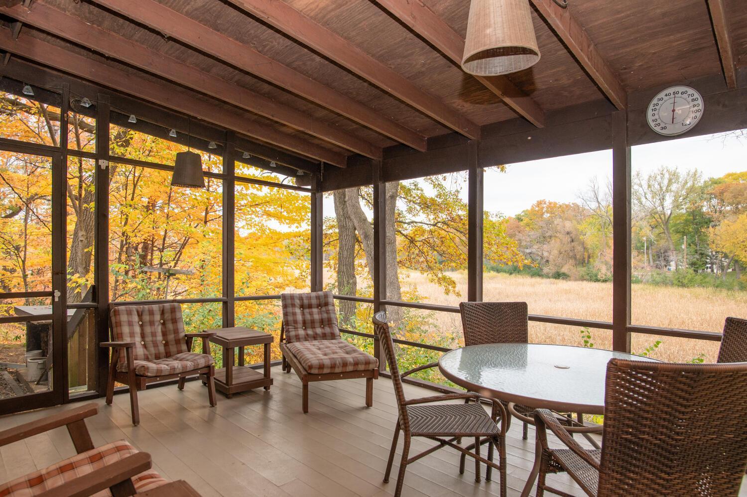 Screened porch with exposed beams and patio seating overlooking autumn trees and meadow.