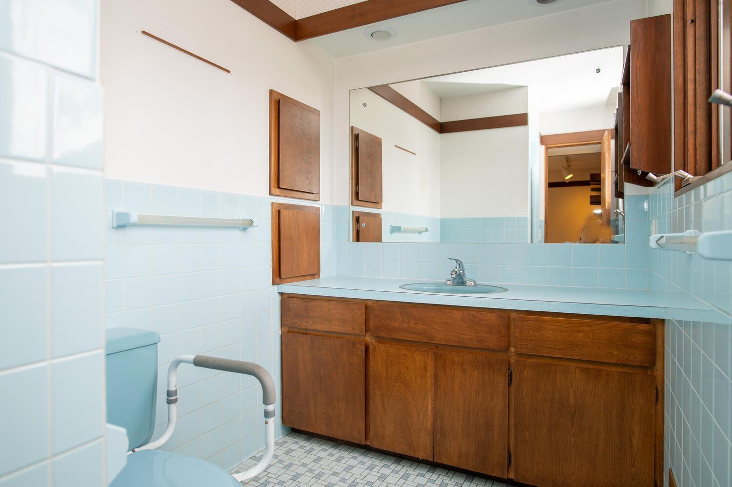 Bathroom with pale blue tile, a large wood vanity, wall mirror, and toilet grab bars.
