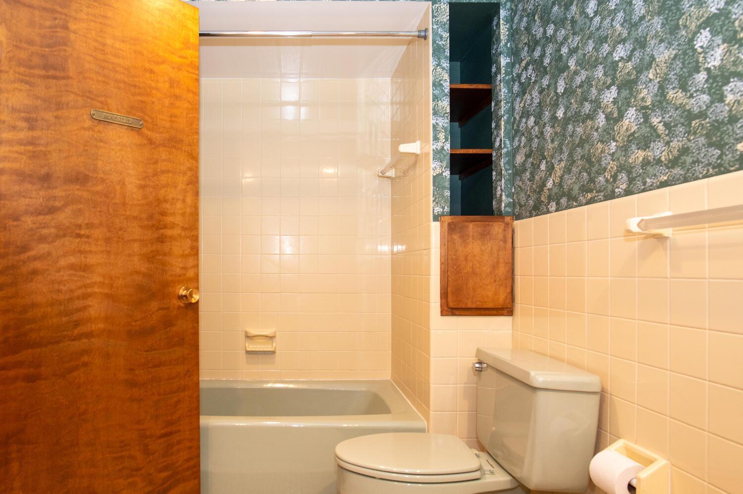 Bathroom with tiled tub and shower, toilet, and dark patterned wallpaper.