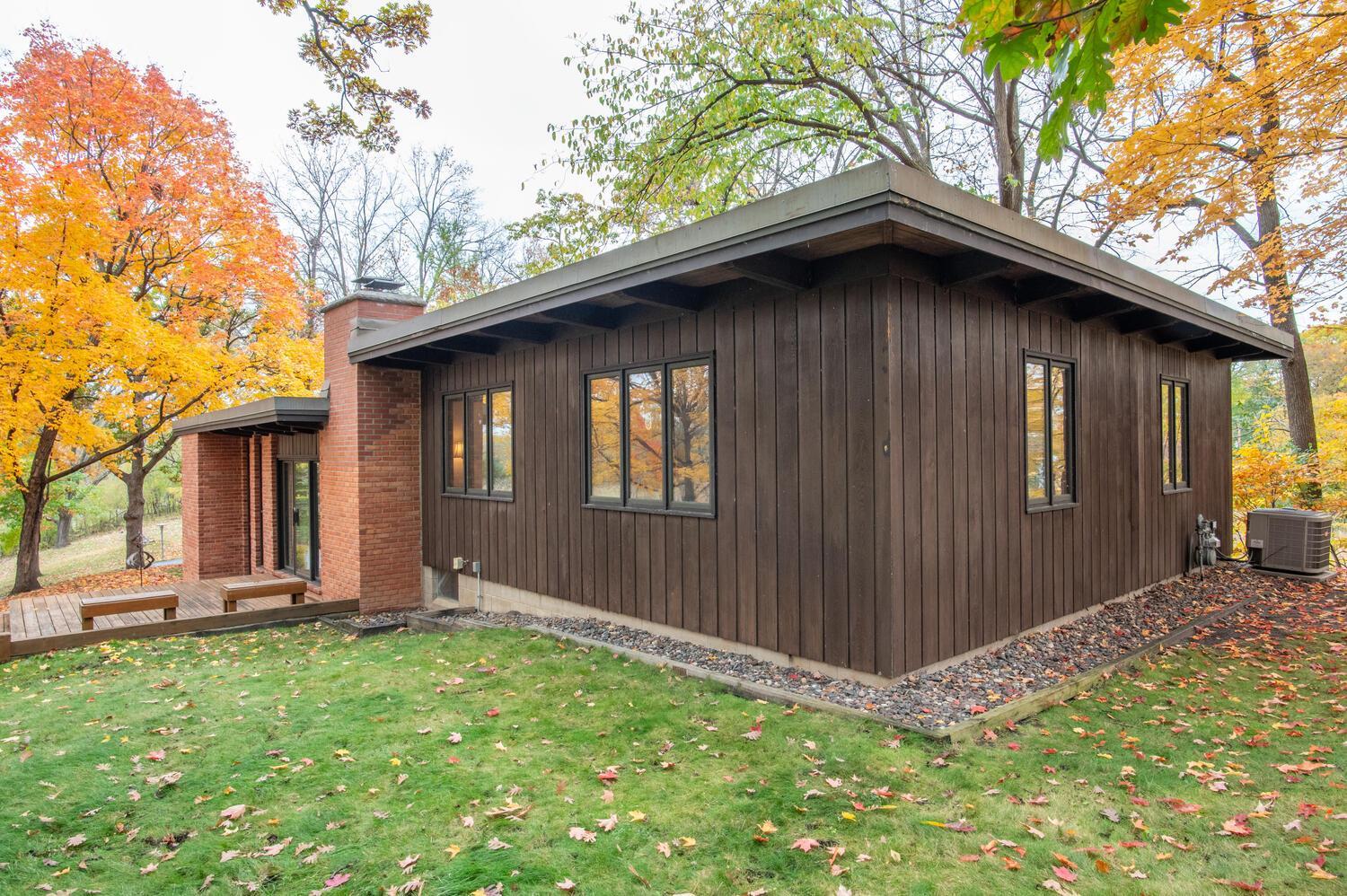 Modern wood-sided home with a brick chimney and small deck, surrounded by autumn trees.