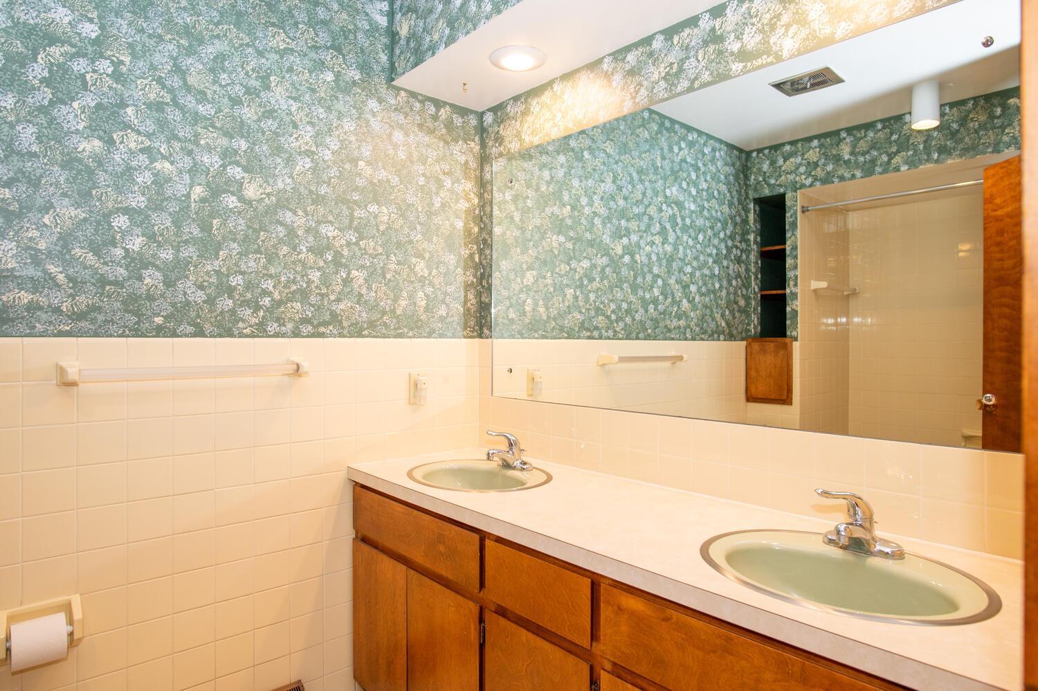 Bathroom with a double-sink wood vanity, large mirror, and teal patterned wallpaper.