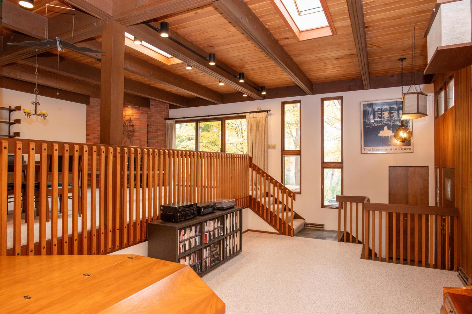 Split-level living area with vaulted wood ceiling, skylights, and railings beside tall windows.