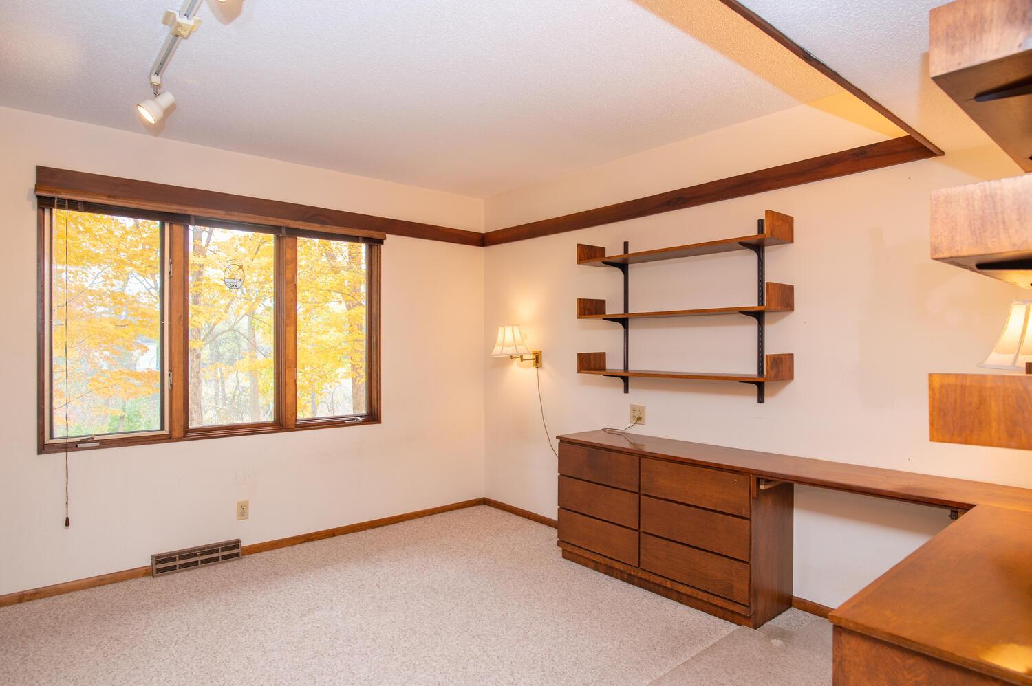 Home office with built-in wooden desk and shelves beside a large window overlooking autumn trees.