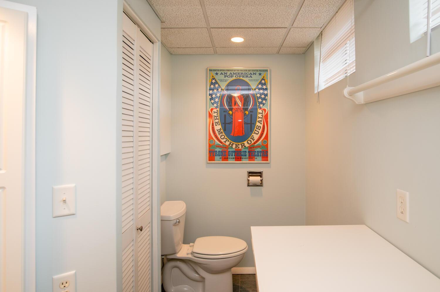 Bathroom with a toilet, wide white counter, louvered doors, and colorful opera poster.