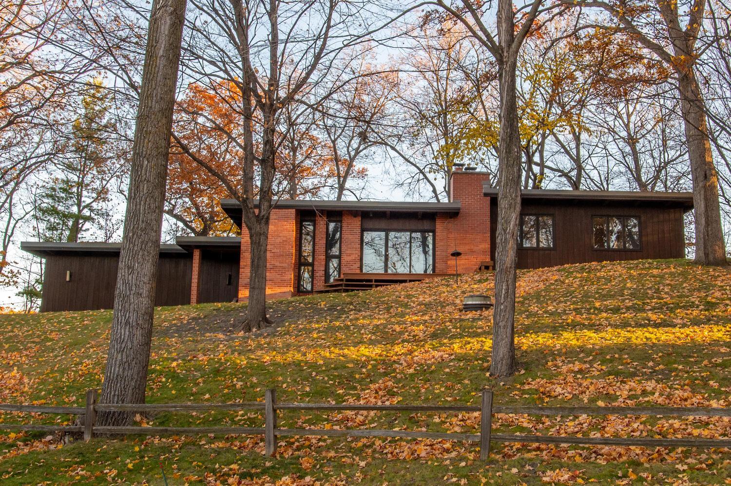 Modern brick and dark wood home among autumn trees on a sloping, leaf-covered lawn.
