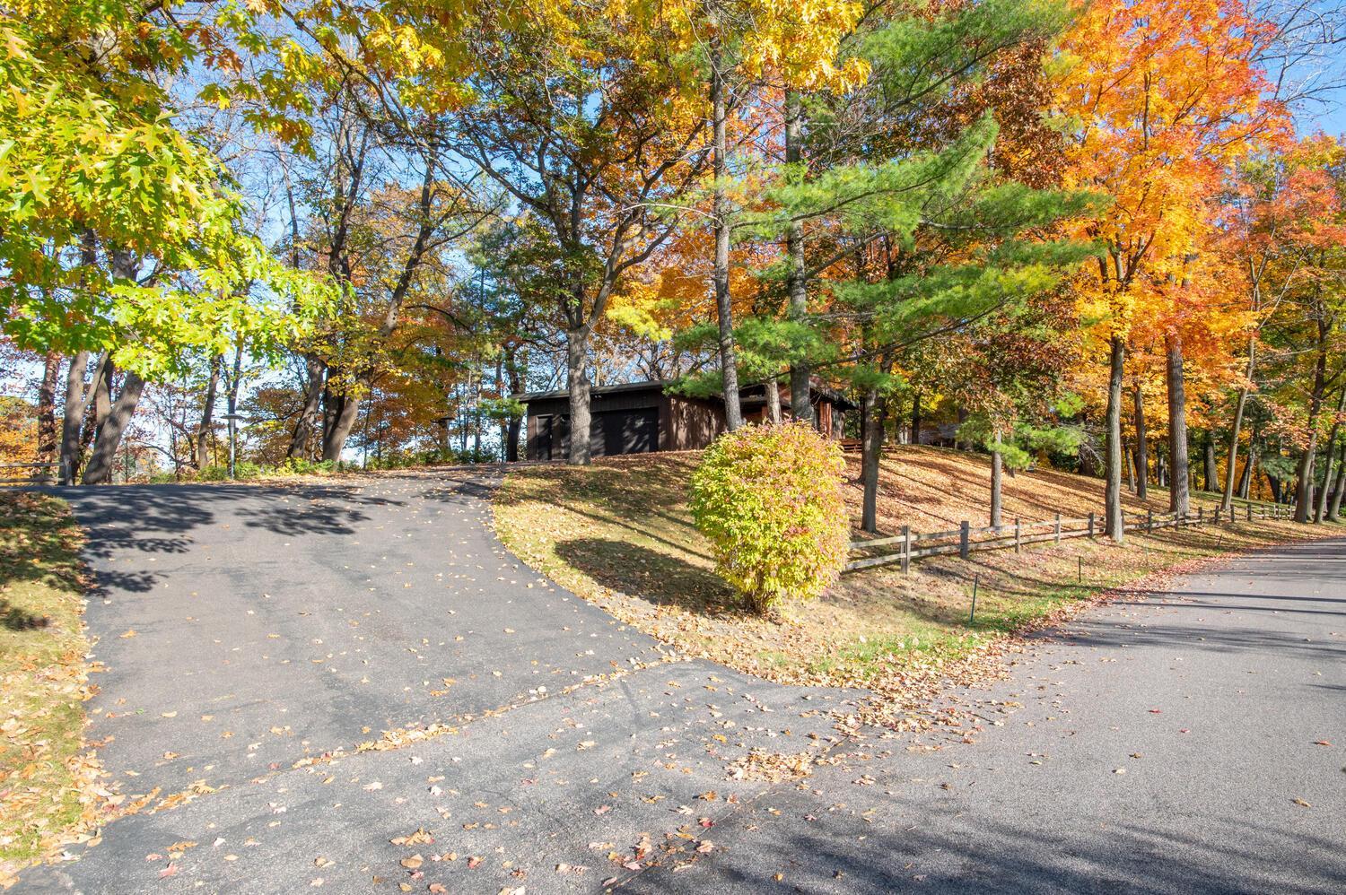 Dark house among colorful autumn trees, approached by a curving driveway and bordered by a low fence.