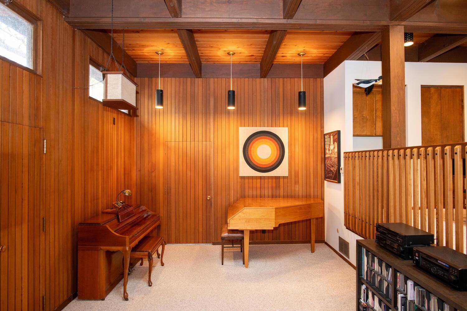 Wood-paneled music room with two pianos, exposed ceiling beams, and a wooden balcony railing.