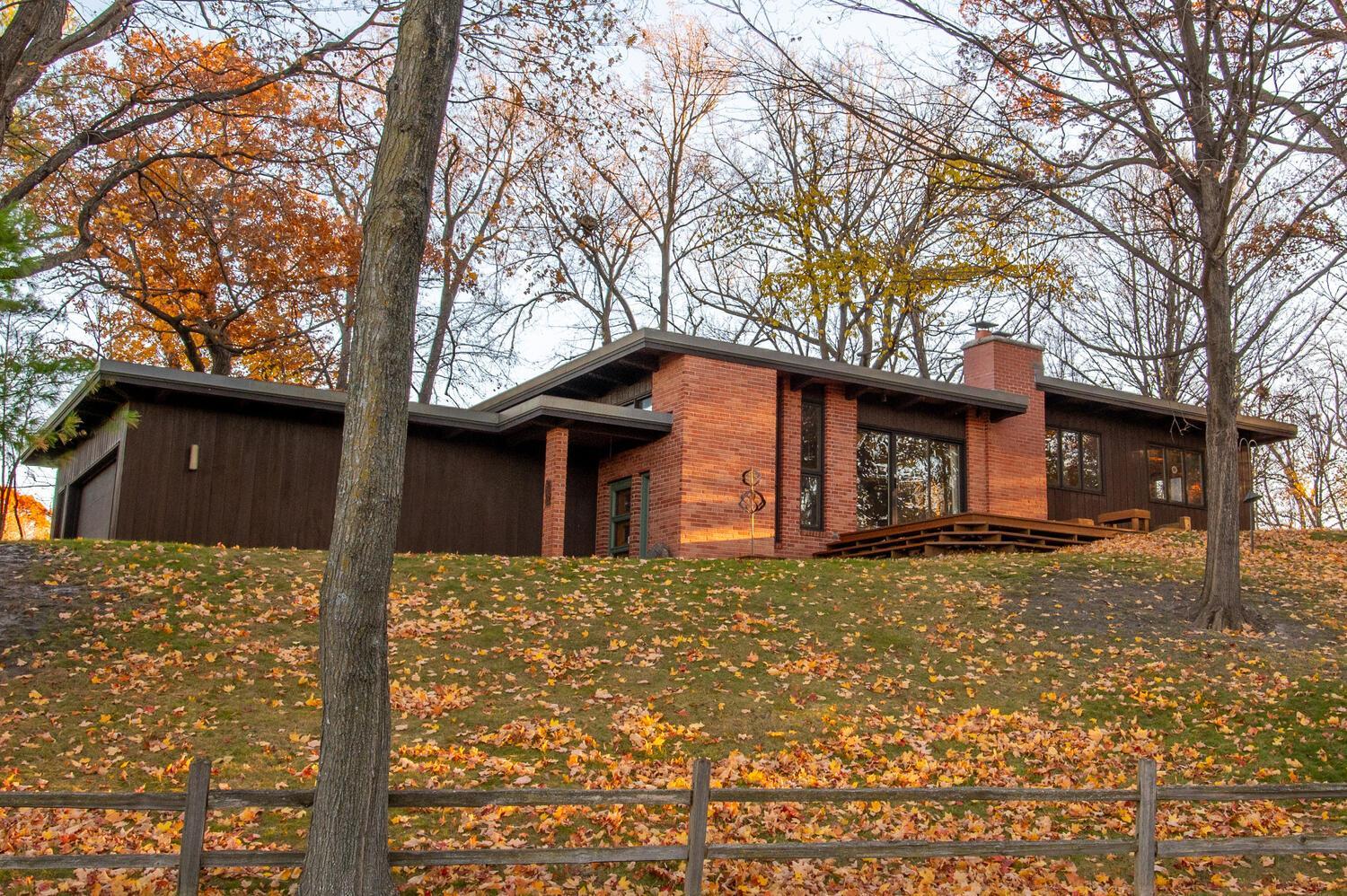 Mid-century modern home with red brick and dark wood siding on a leafy autumn slope.